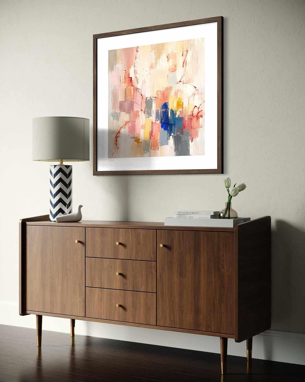 A framed abstract art print with a white mat and dark wood frame hangs on a light beige wall above a mid-century modern wooden sideboard. The room is styled with a gray lamp, a small decorative bird figurine, and a vase with tulips.