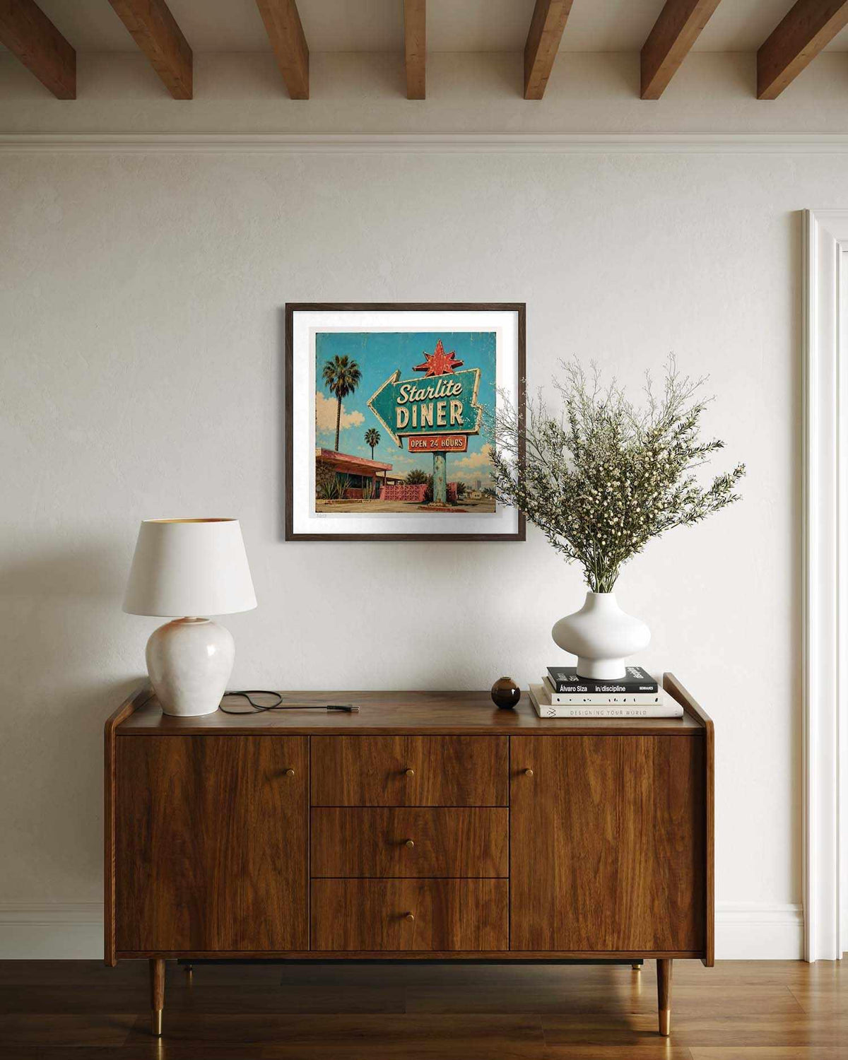 A framed retro-style 'Smile Diner' art print hangs on a cream wall above a mid-century modern wooden sideboard. The room is styled with a white ceramic table lamp, a white vase of dried branches, and books.