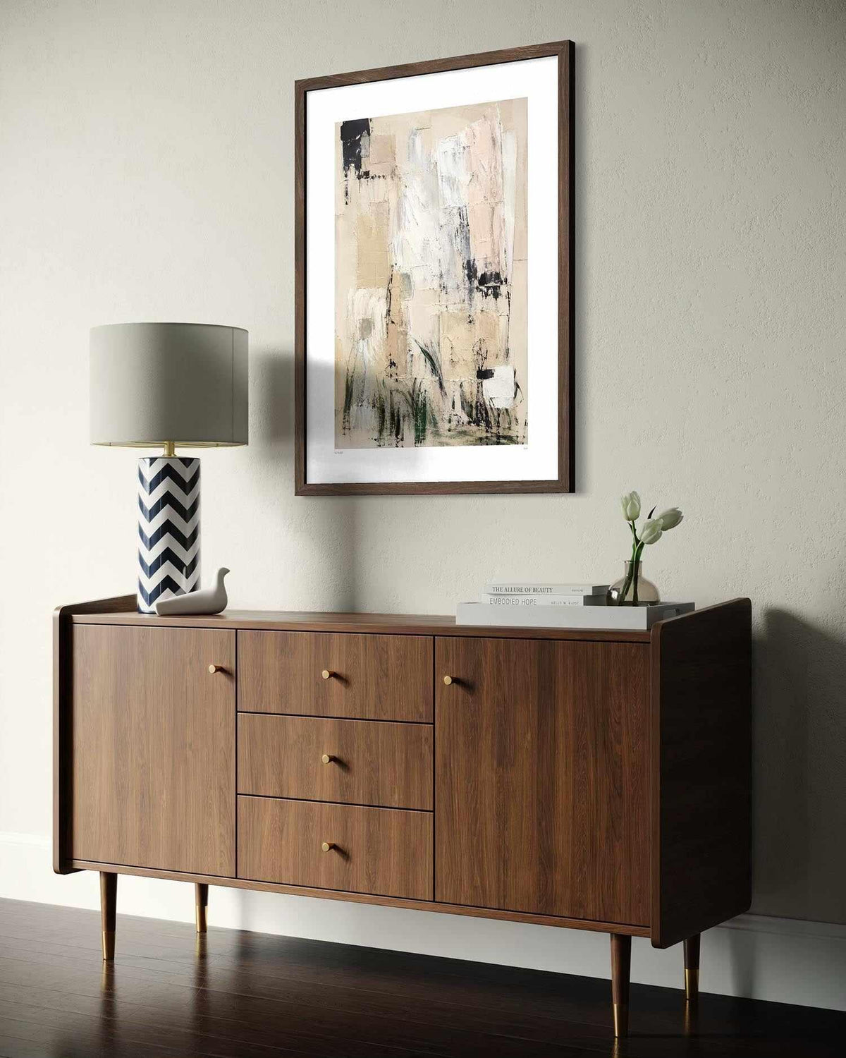 A mid-century modern wooden sideboard cabinet with gold-tone knobs, styled in a room setting with a framed abstract print on the wall, a patterned lamp, and decorative items.