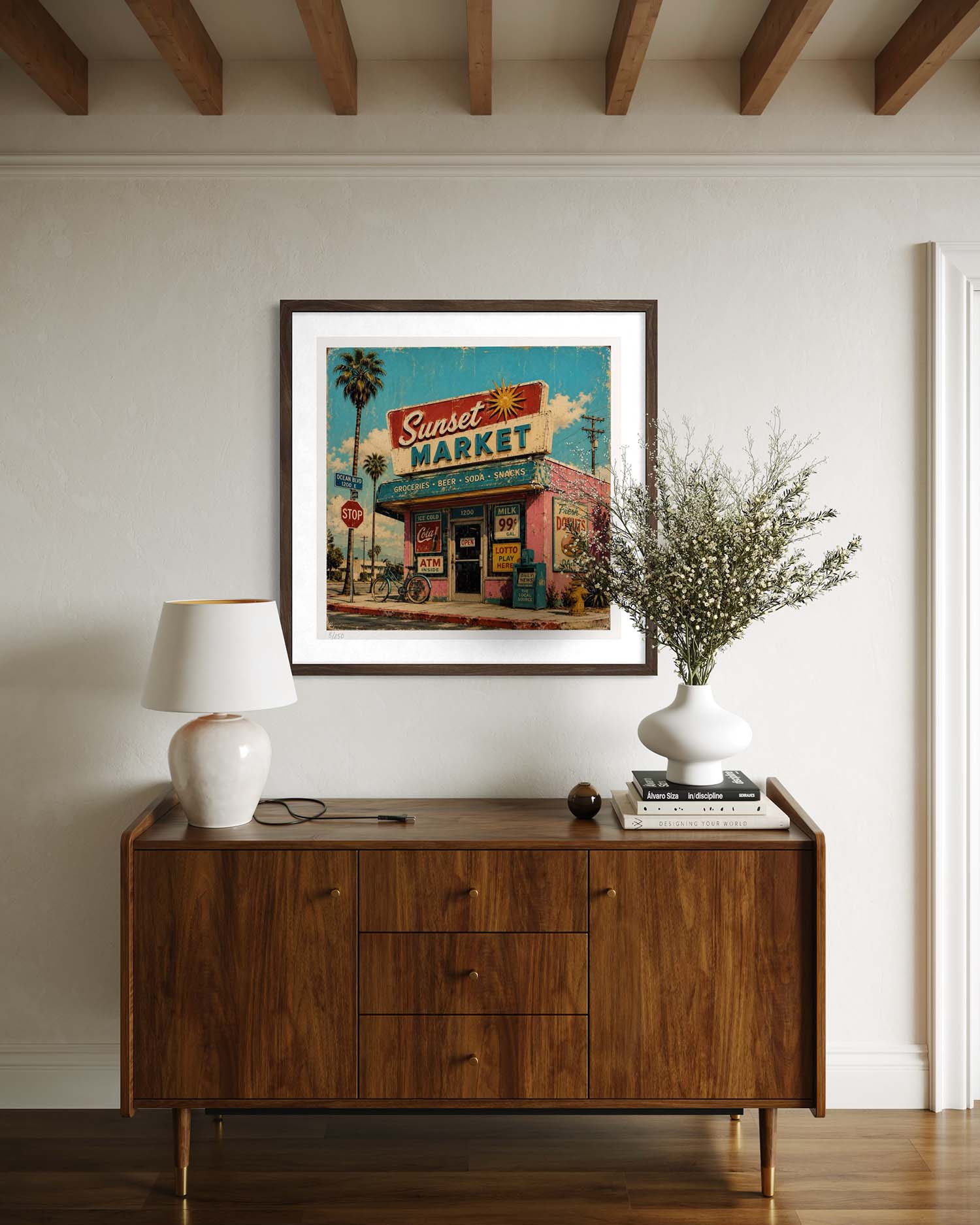 A framed art print of a retro 'Sunset Market' sign hanging on a beige wall above a mid-century modern wooden sideboard. The print features a colorful illustration with a blue sky, palm tree, and warm-toned storefront, styled in a home interior with a white lamp and a vase of branches.