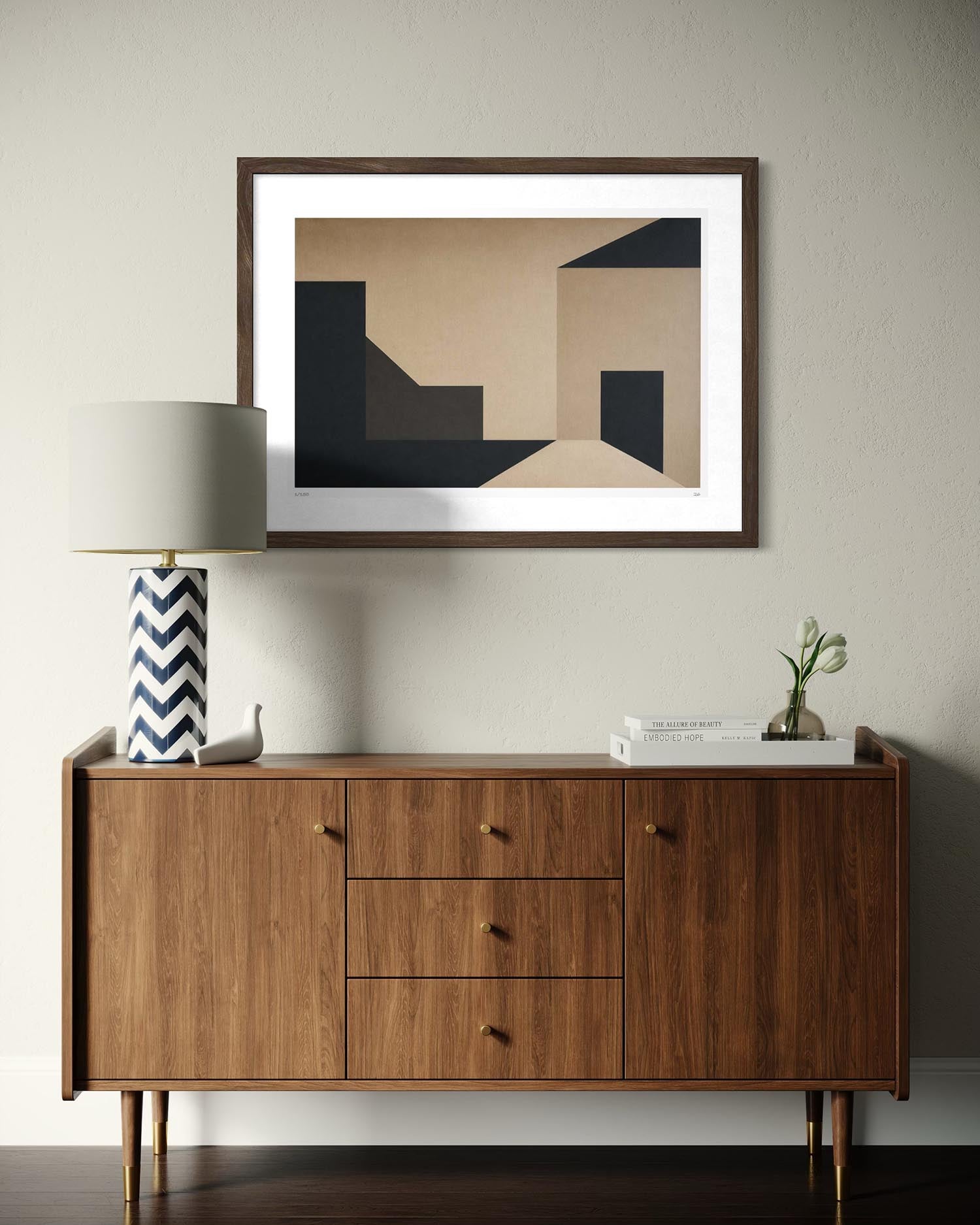 A framed abstract geometric art print with beige, black, and brown shapes, displayed on a light gray wall above a mid-century modern wooden sideboard with a lamp and books.