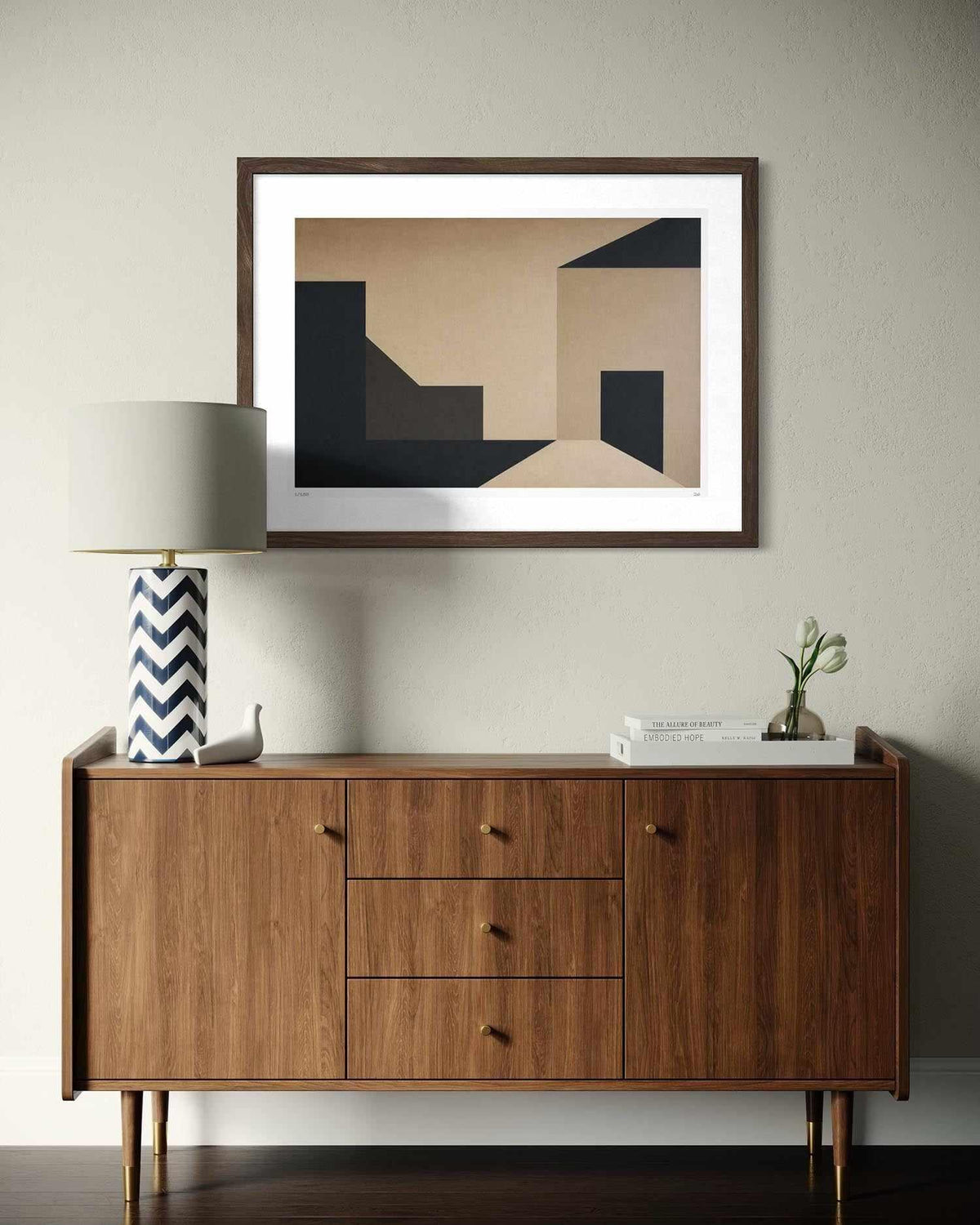 A framed abstract geometric art print with beige, black, and brown shapes, displayed on a light gray wall above a mid-century modern wooden sideboard with a lamp and books.