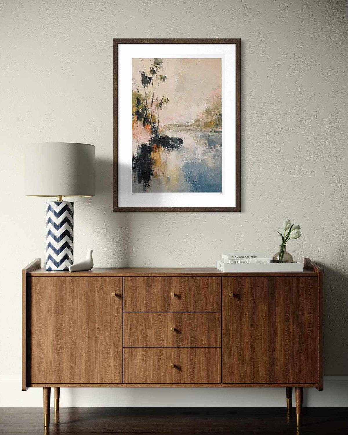 A framed abstract landscape painting in a dark wood frame hangs on a light gray wall above a mid-century modern wooden sideboard with a lamp and decorative items.