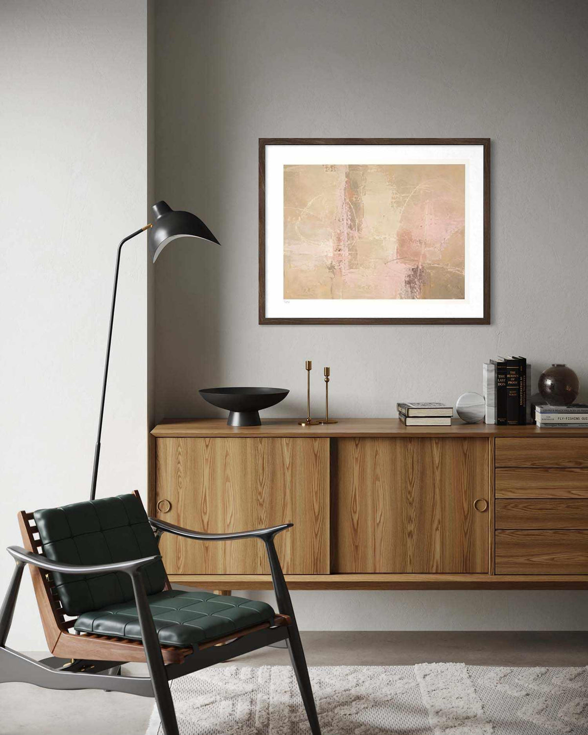 A framed abstract art print with soft beige, pink, and brown tones hangs on a gray wall above a mid-century modern wooden credenza in a styled living room setting.