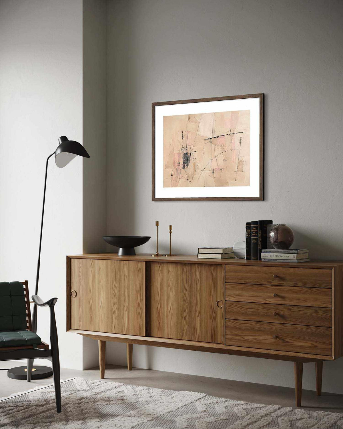 A framed abstract art print with a white mat and dark wood frame hangs on a gray wall above a mid-century modern wooden sideboard in a styled living room setting.