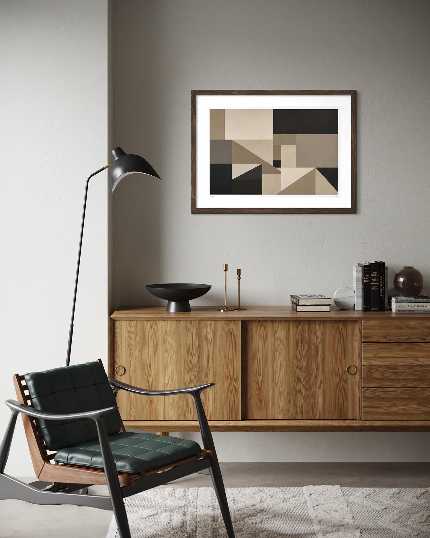 A framed abstract geometric art print with beige, black, and brown shapes hangs on a gray wall above a mid-century modern wooden credenza in a styled living room setting.