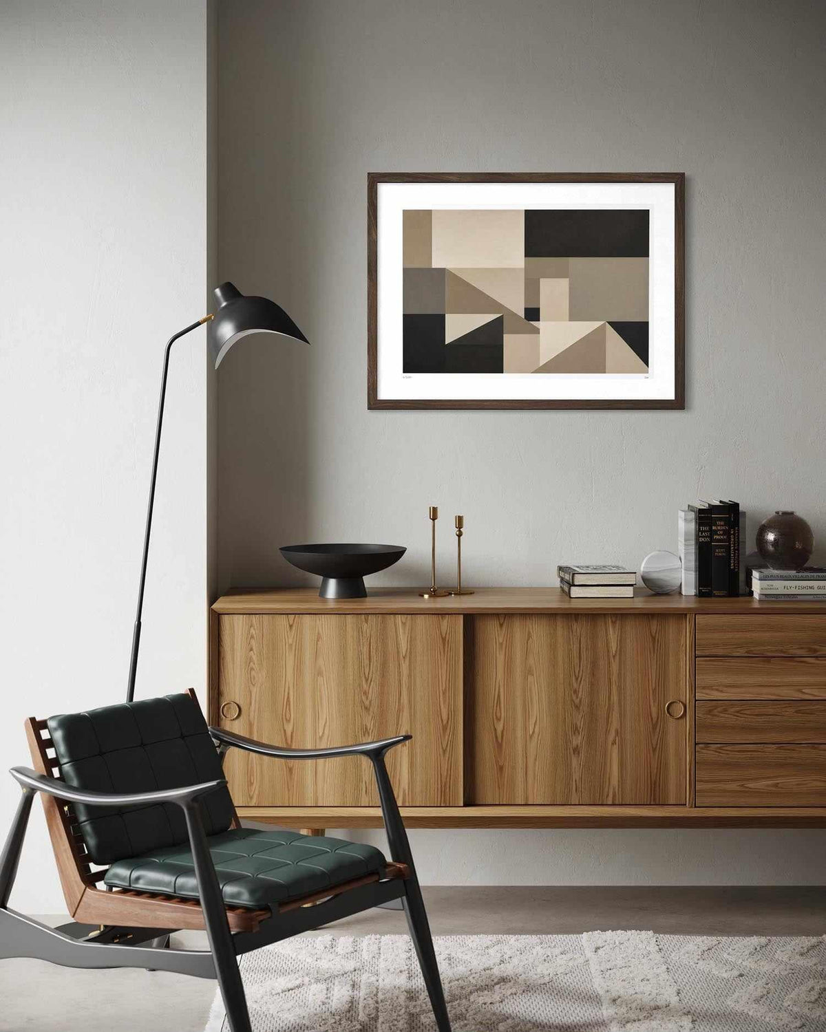 A framed abstract geometric art print with beige, black, and brown shapes hangs on a gray wall above a mid-century modern wooden credenza in a styled living room setting.
