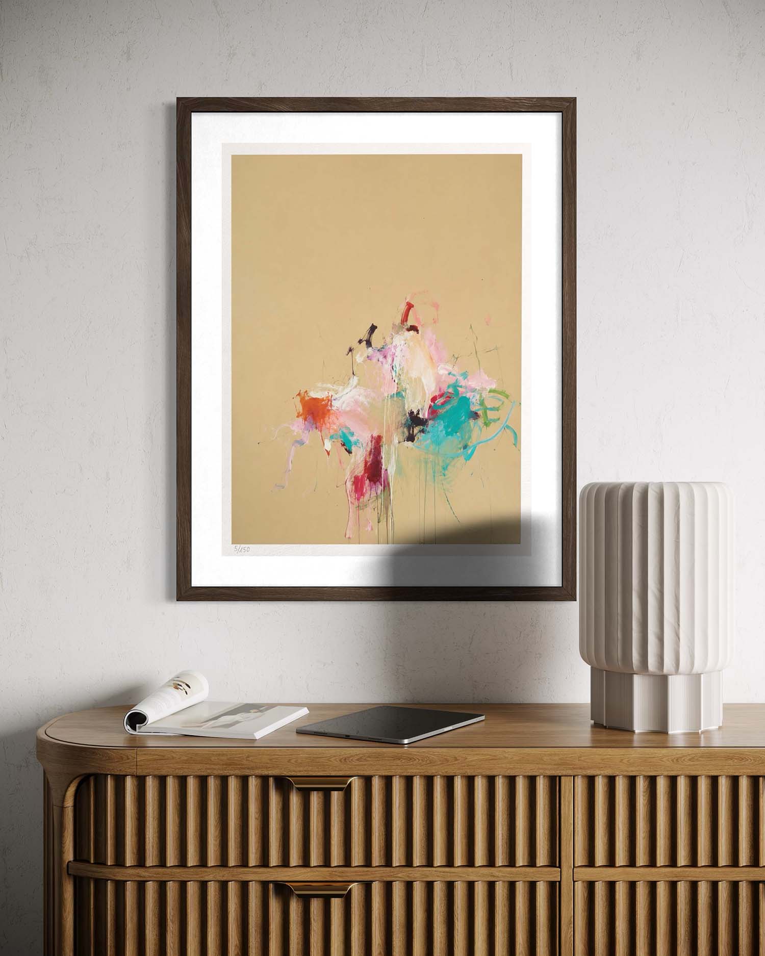 A framed abstract art print featuring colorful paint splatters on a warm beige background, styled on a light gray wall above a wooden cabinet with a lamp and magazine nearby.