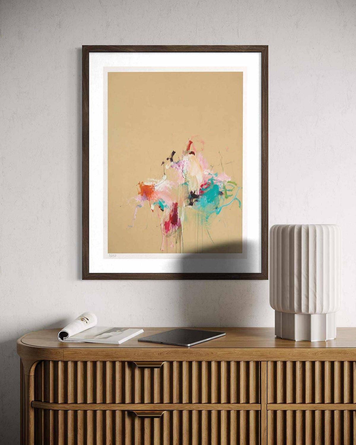 A framed abstract art print featuring colorful paint splatters on a warm beige background, styled on a light gray wall above a wooden cabinet with a lamp and magazine nearby.