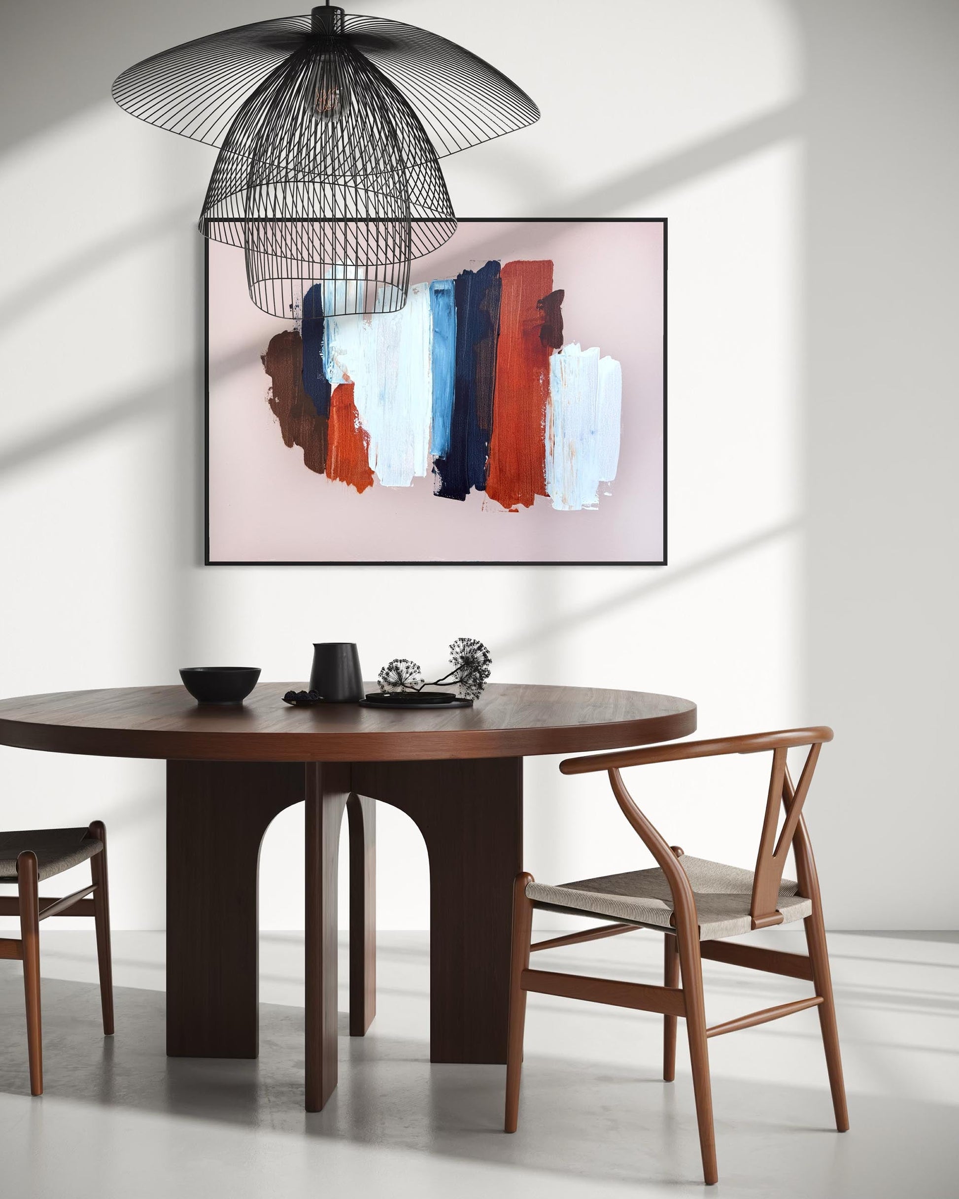 A modern dining room scene featuring a round dark wood table, two wooden chairs, a black wire pendant lamp, and a framed abstract wall art print with vertical brushstrokes in brown, orange, blue, white, and pink on a light wall.