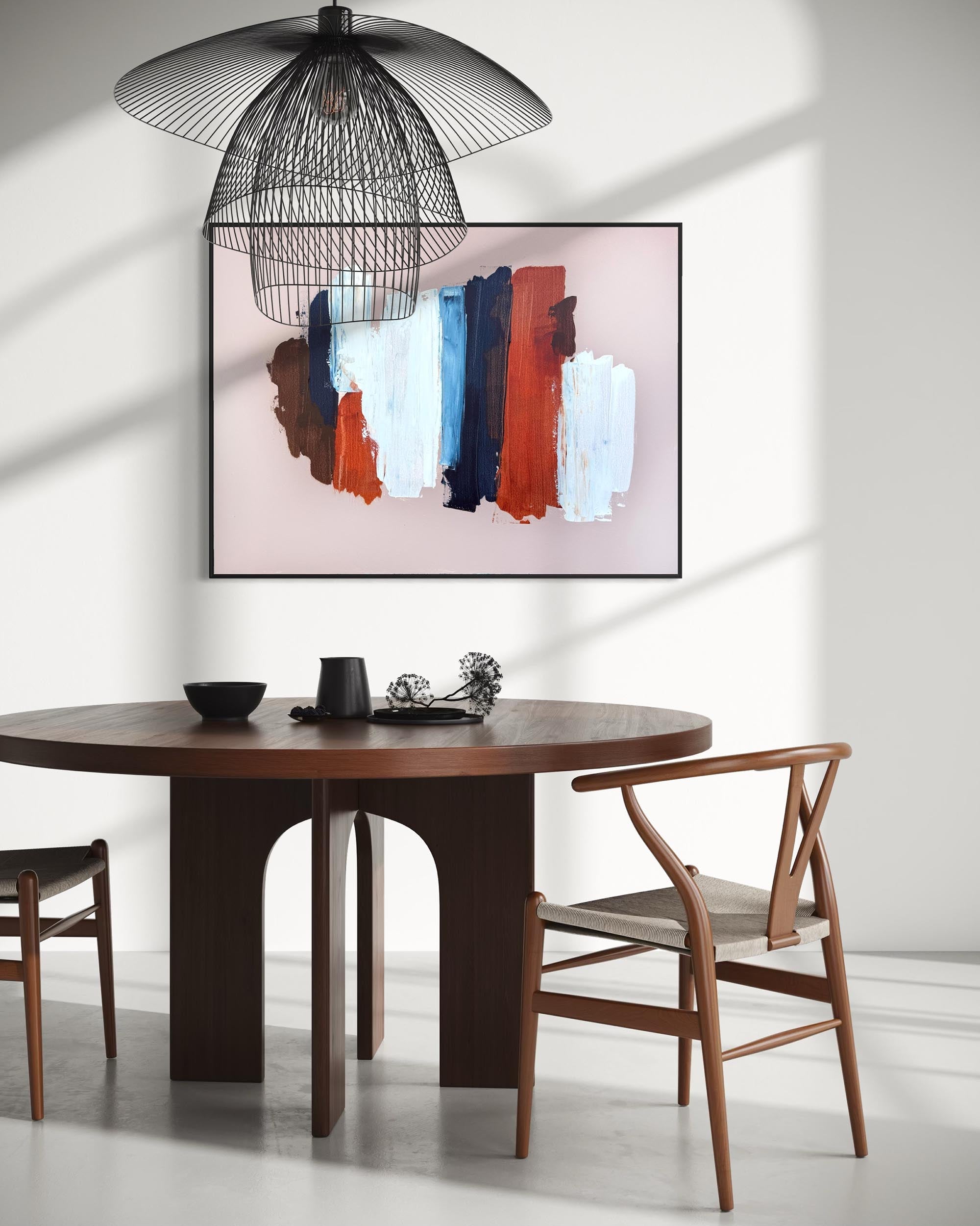 A modern dining room scene featuring a round dark wood table, two wooden chairs, a black wire pendant lamp, and a framed abstract wall art print with vertical brushstrokes in brown, orange, blue, white, and pink on a light wall.