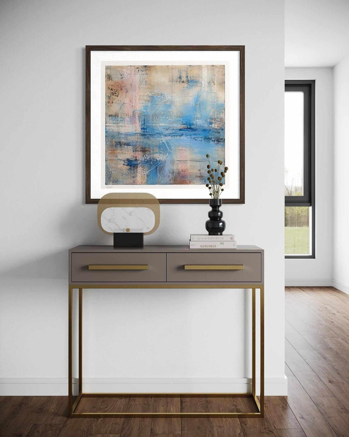 A framed abstract painting with blue, beige, and pink tones hangs on a white wall above a modern console table with gold-tone legs and handles. The room has wood flooring and a window in the background.