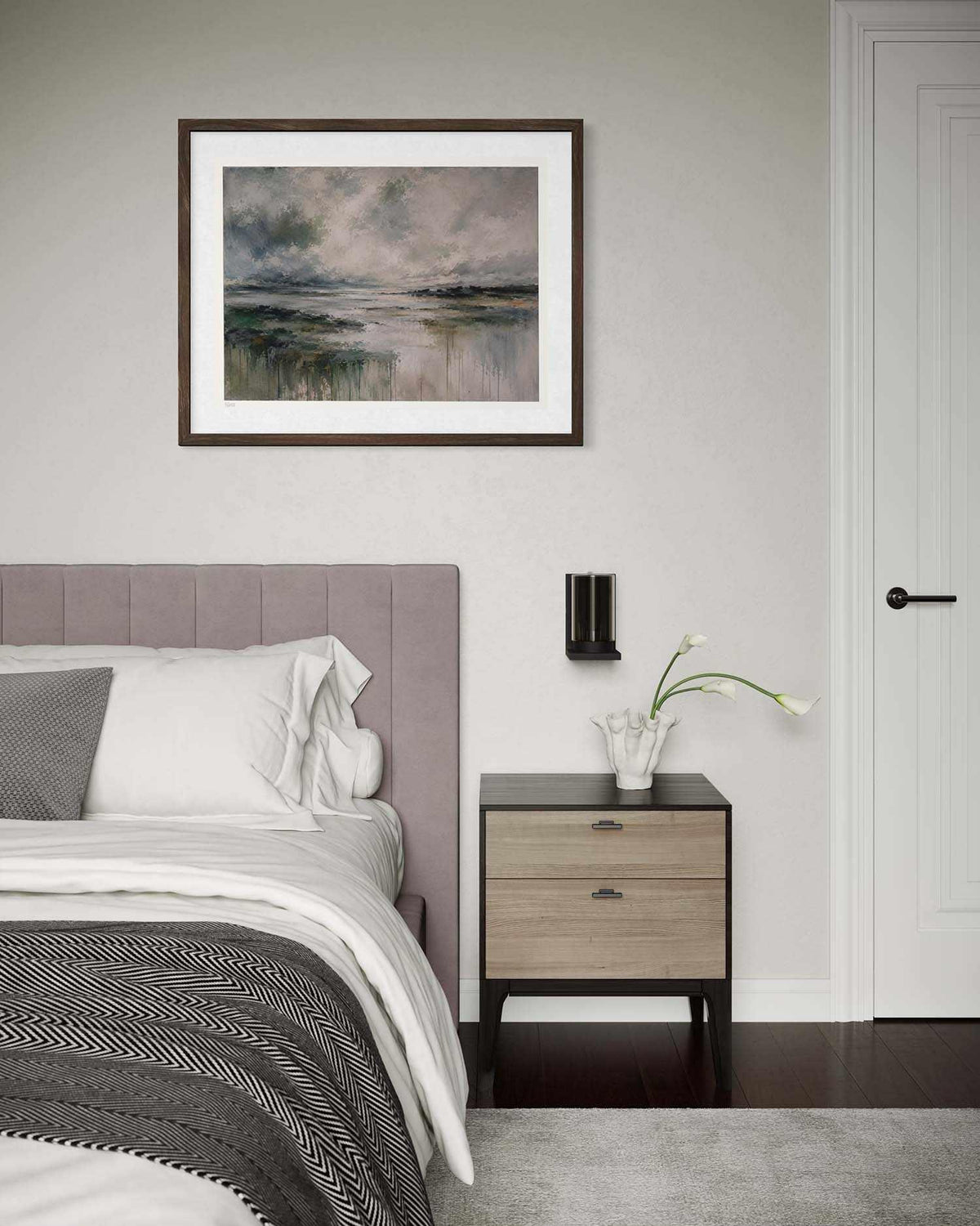 A framed landscape art print hangs on a light gray wall above a bed in a modern bedroom setting. The artwork depicts a misty, moody lake or river scene with muted blue, gray, and green tones, surrounded by a white mat and dark brown frame.