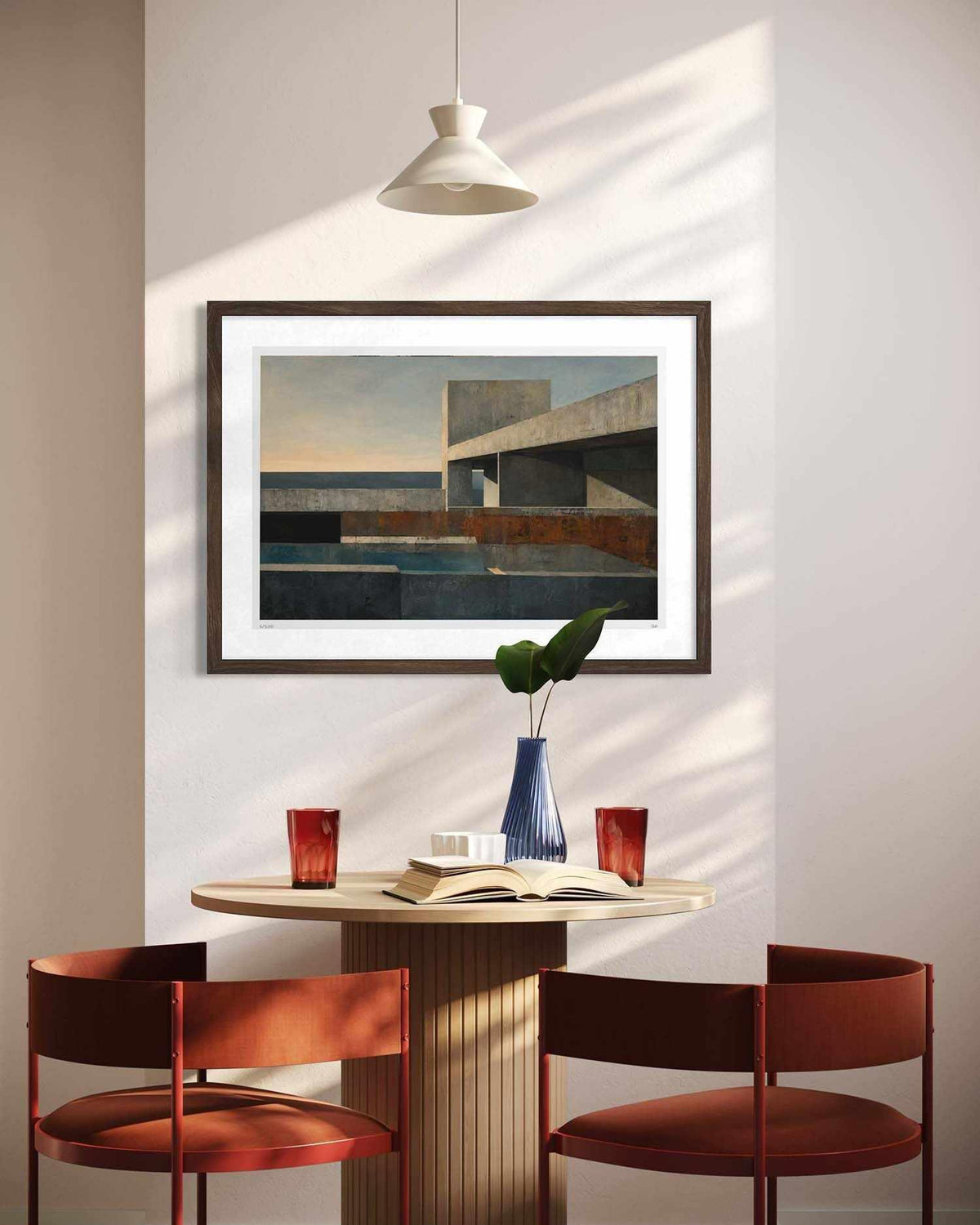 A framed abstract architectural painting hangs on a beige wall above a small round table with two rust-red chairs, a blue vase with green leaves, and red glass cups. A white pendant light casts diagonal shadows across the wall and table.