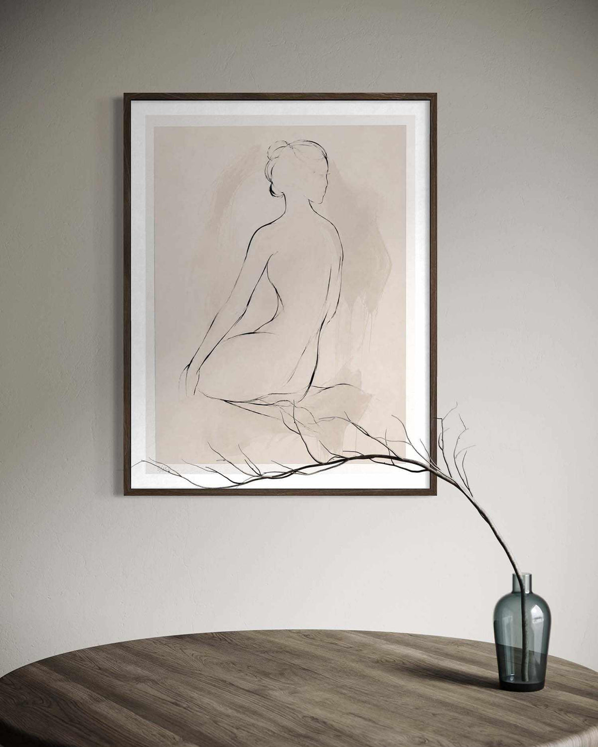 A framed minimalist line drawing of a seated nude figure on a beige background, hanging on a light gray wall above a wooden table with a small blue-gray vase holding a branch.