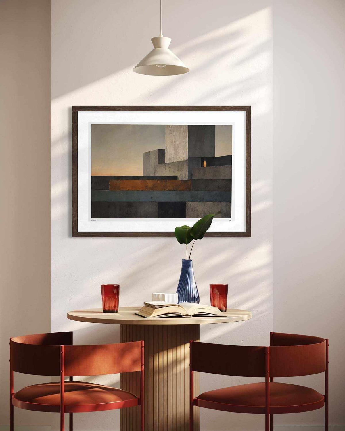A framed abstract geometric painting hangs on a beige wall above a small round table with two rust-red chairs, a blue vase with green leaves, and a hanging pendant light casting shadows.