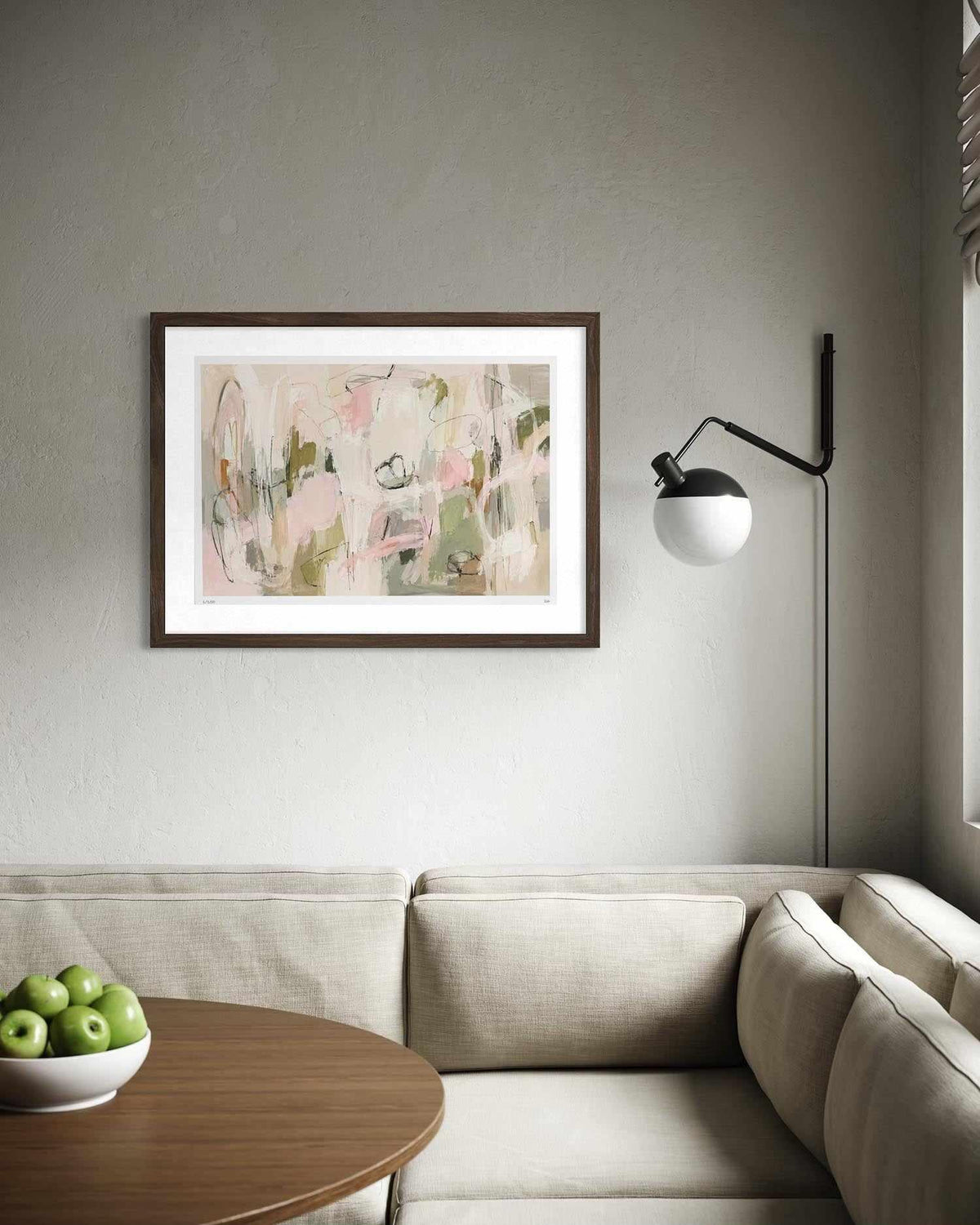 A framed abstract art print with soft pink, beige, and green brushstrokes hangs on a textured gray wall above a light beige sofa. A round wooden coffee table with a bowl of green apples is in the foreground, and a black wall lamp stands to the right.