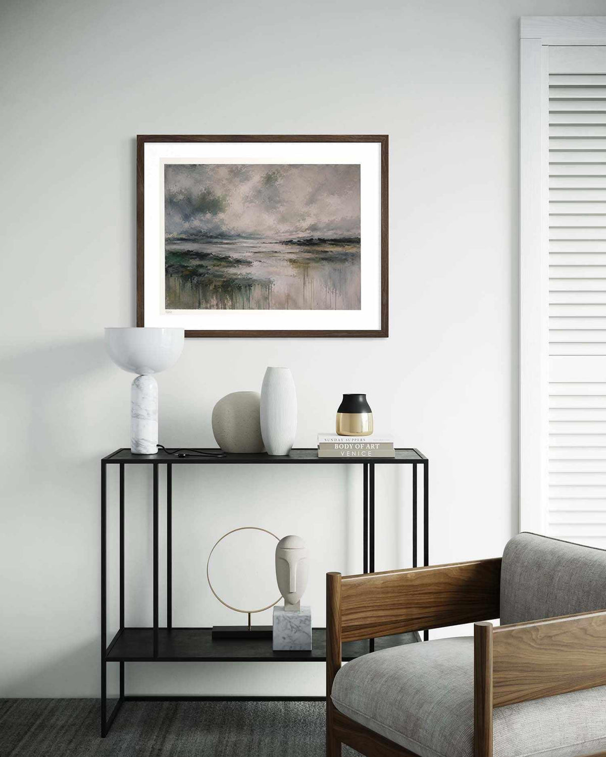 A framed landscape art print hangs on a light gray wall above a black metal console table styled with white and beige ceramic vases and a small lamp. The print depicts a misty, reflective water scene in muted gray, green, and blue tones.