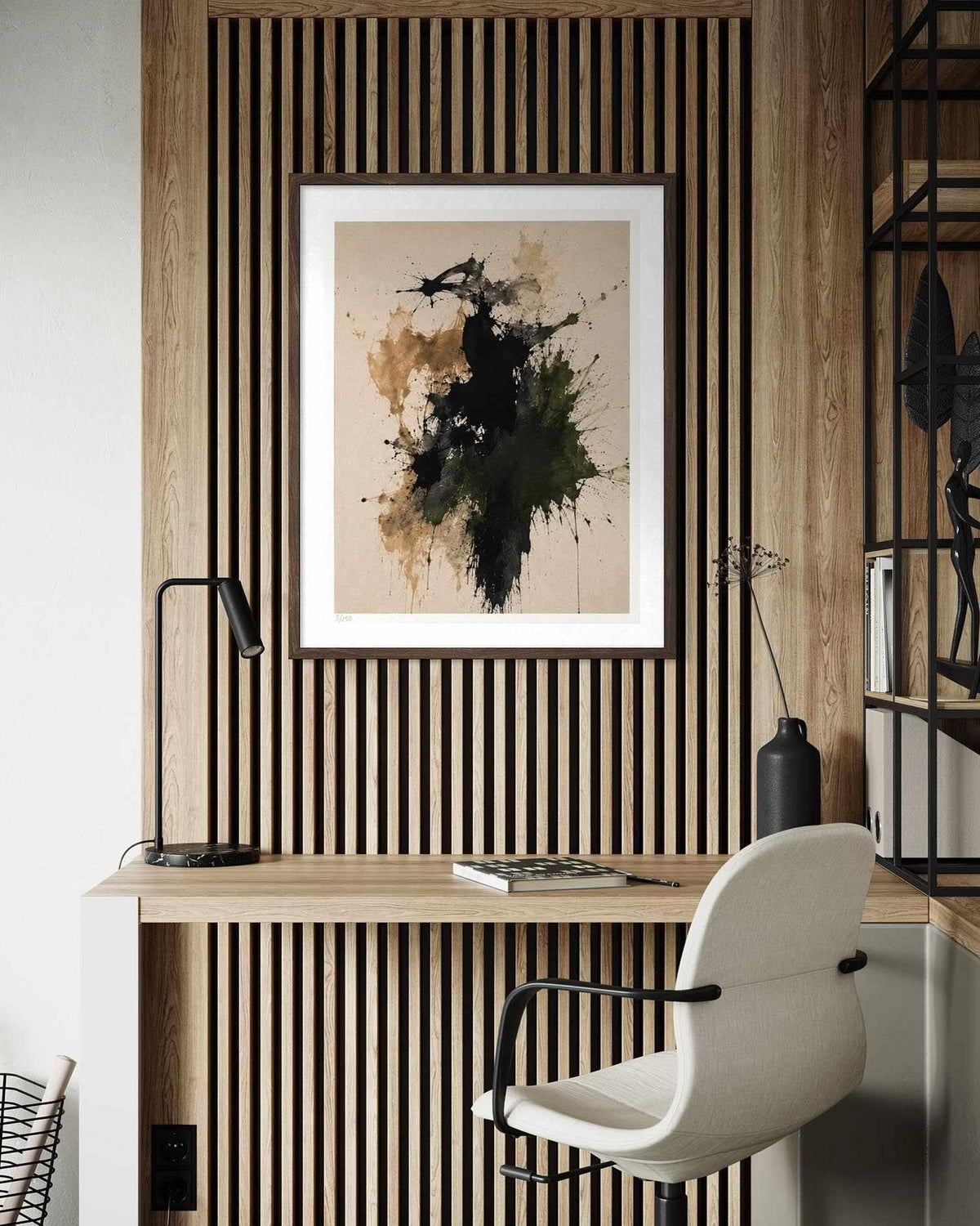 A framed abstract art print with black, green, and brown paint splatters on a beige background, hanging on a wood slat wall above a desk with a white chair in a modern home office setting.