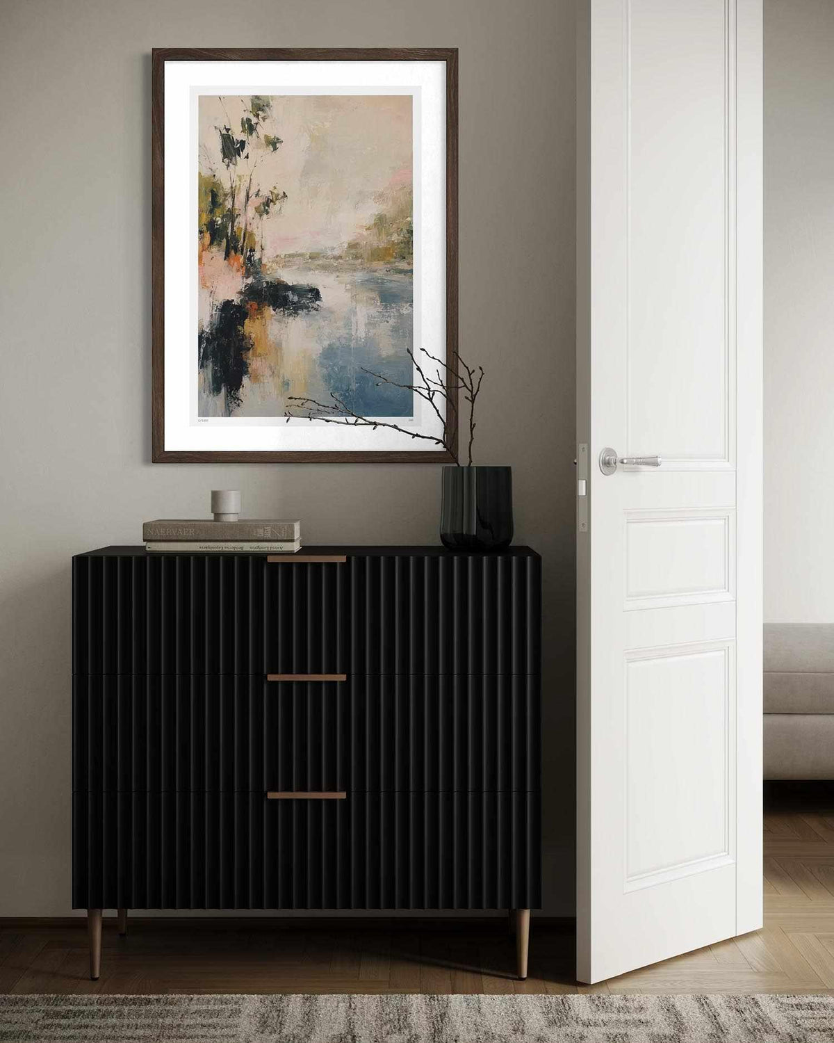 A framed abstract landscape art print hangs on a gray wall above a black ribbed chest of drawers in a modern, styled room.