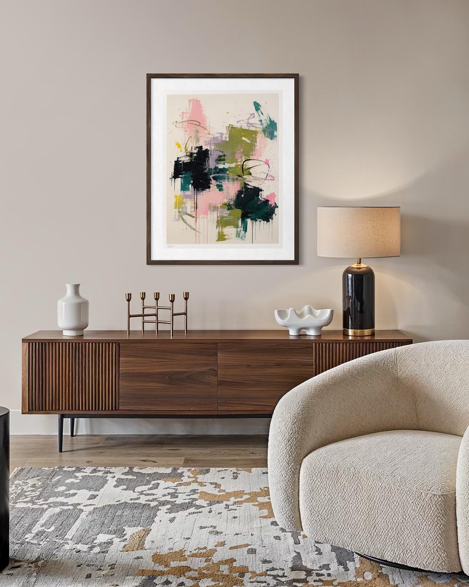 A framed abstract art print with pink, green, black, and beige brushstrokes hangs on a beige wall above a mid-century modern wooden sideboard in a styled living room setting.