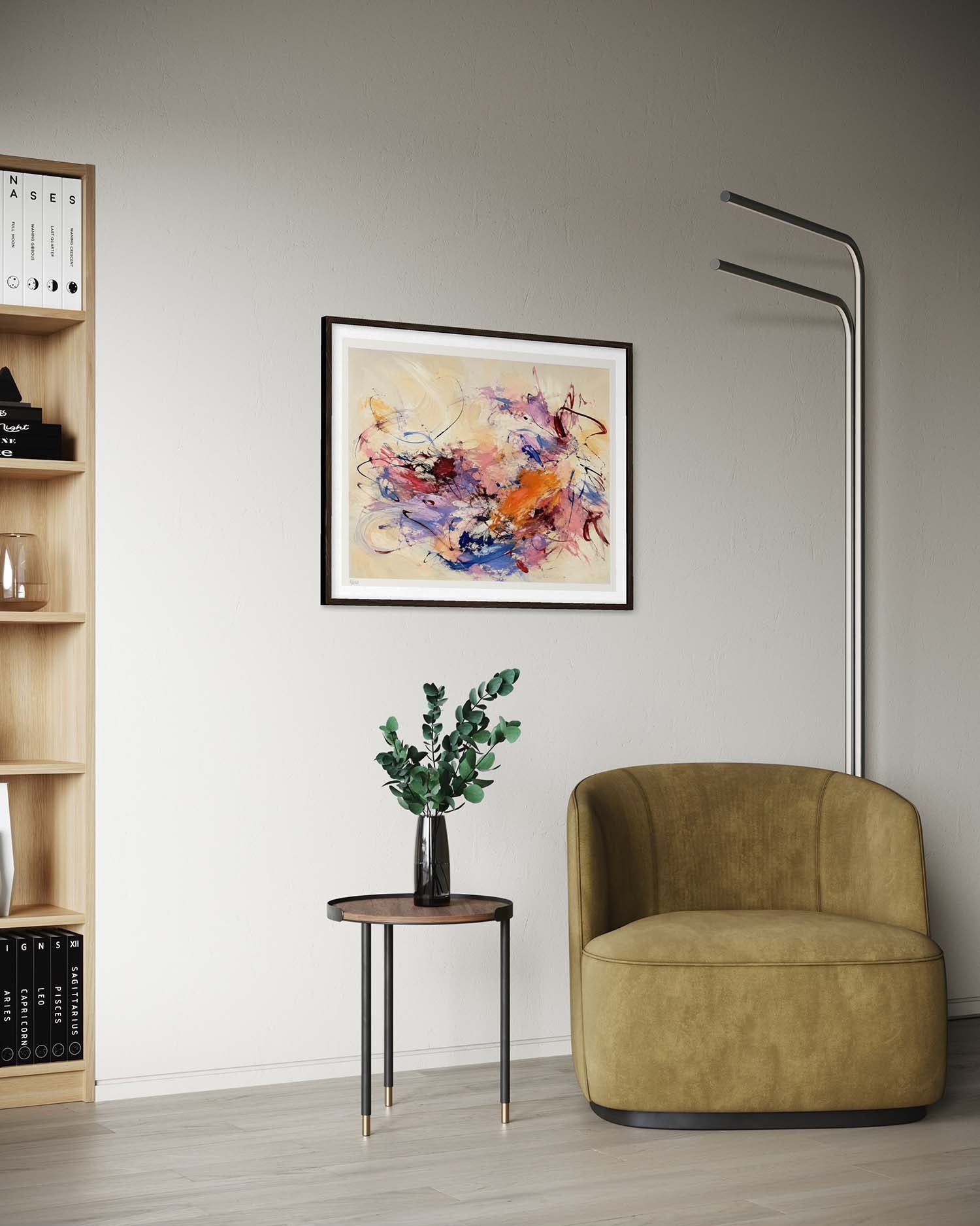 A framed abstract painting with vibrant splashes of orange, purple, blue, and pink on a cream background, displayed on a light gray wall in a modern living room setting with a mustard green armchair, a small side table with a plant, and a wooden bookshelf.