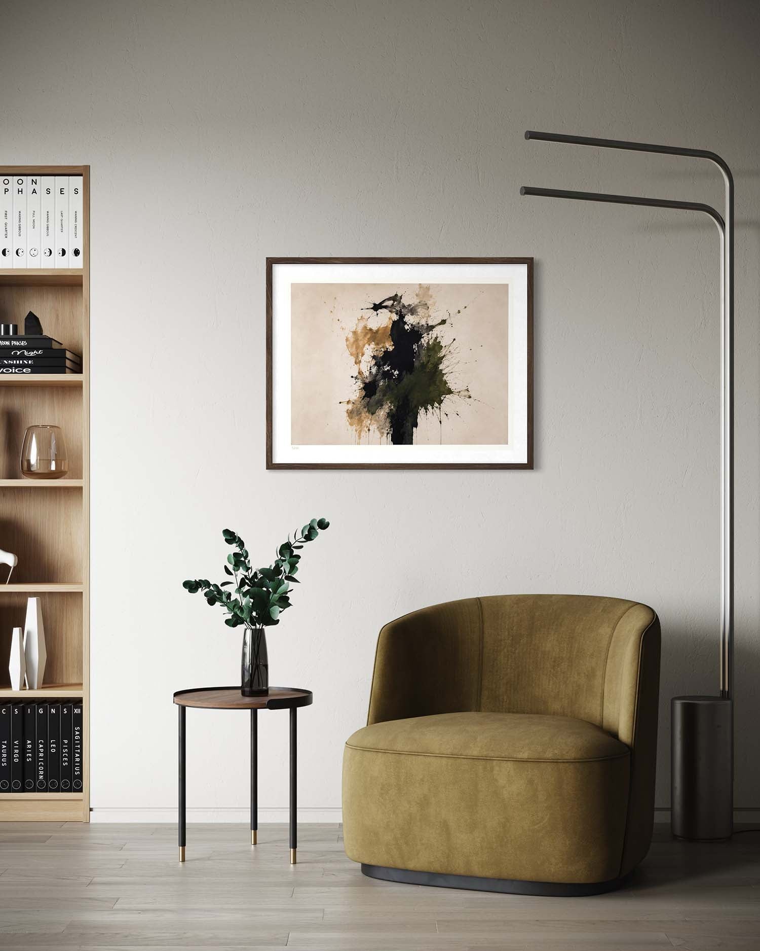 A framed abstract art print featuring black, brown, and gold paint splatters on a beige background, styled in a modern living room setting above a green armchair.