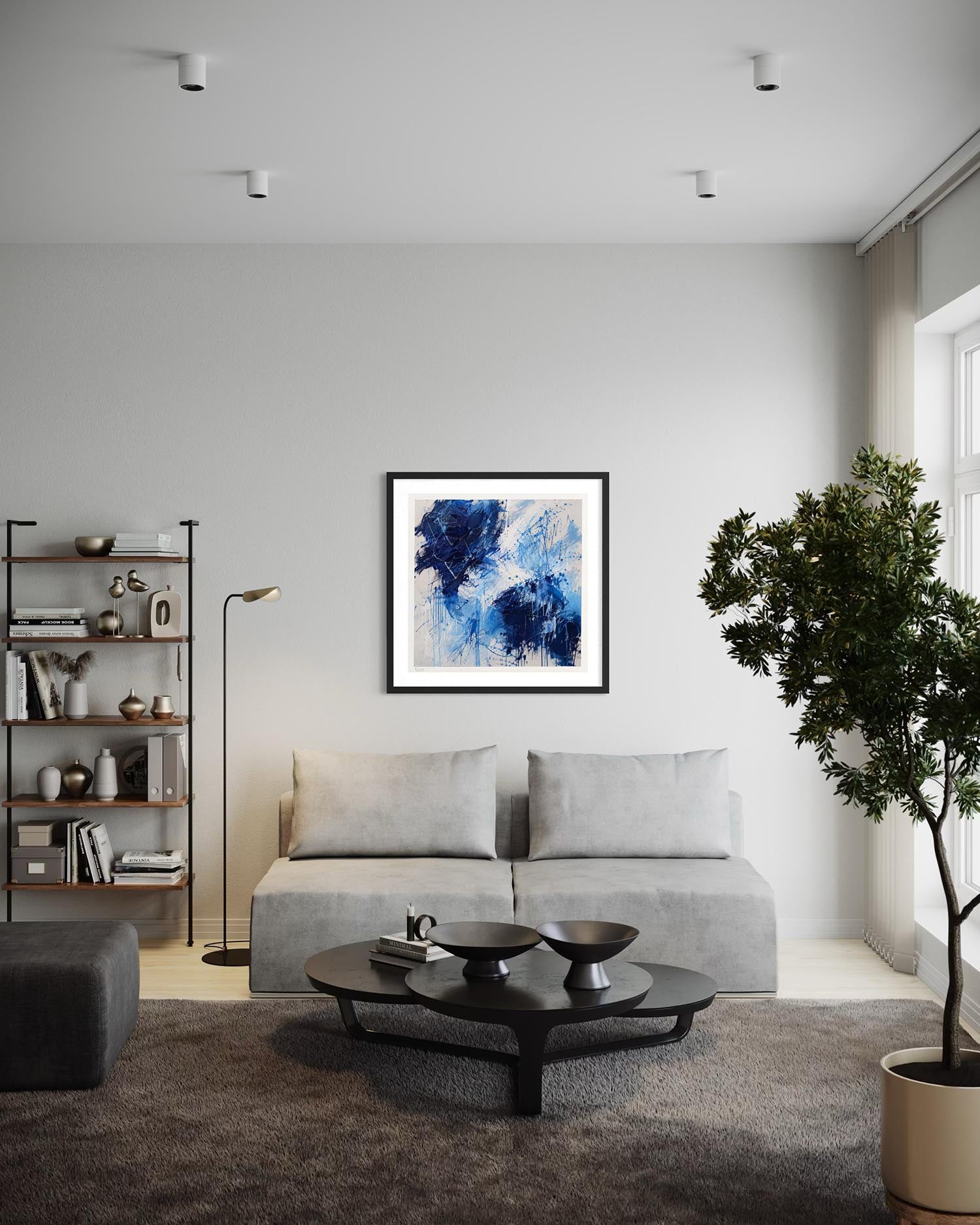 A framed abstract painting with blue, navy, and white brushstrokes hangs on a light gray wall above a modern light gray sofa in a styled living room setting.