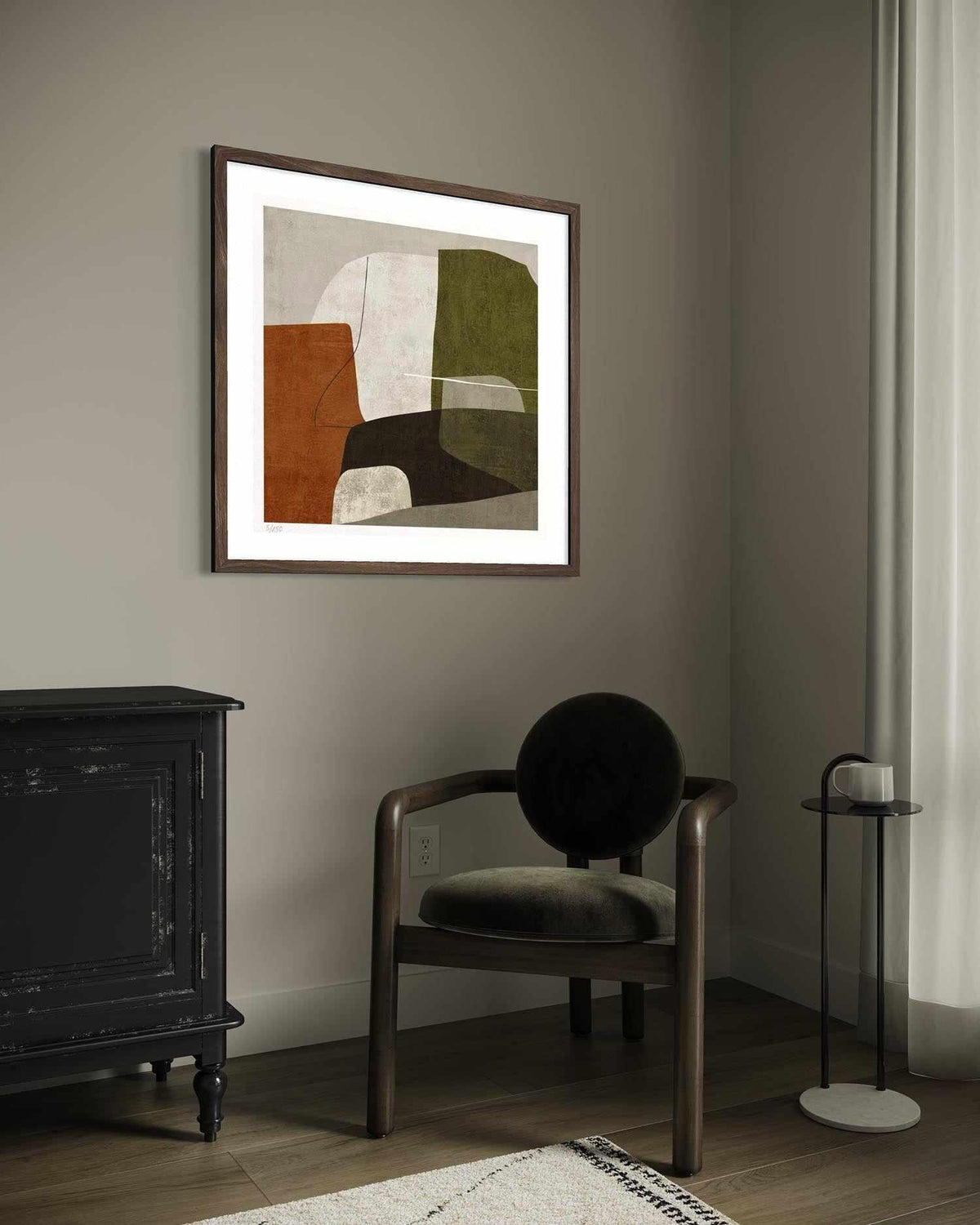 A framed abstract art print hangs on a taupe-gray wall in a styled room setting, featuring a dark wood armchair, a black cabinet, and a small side table.