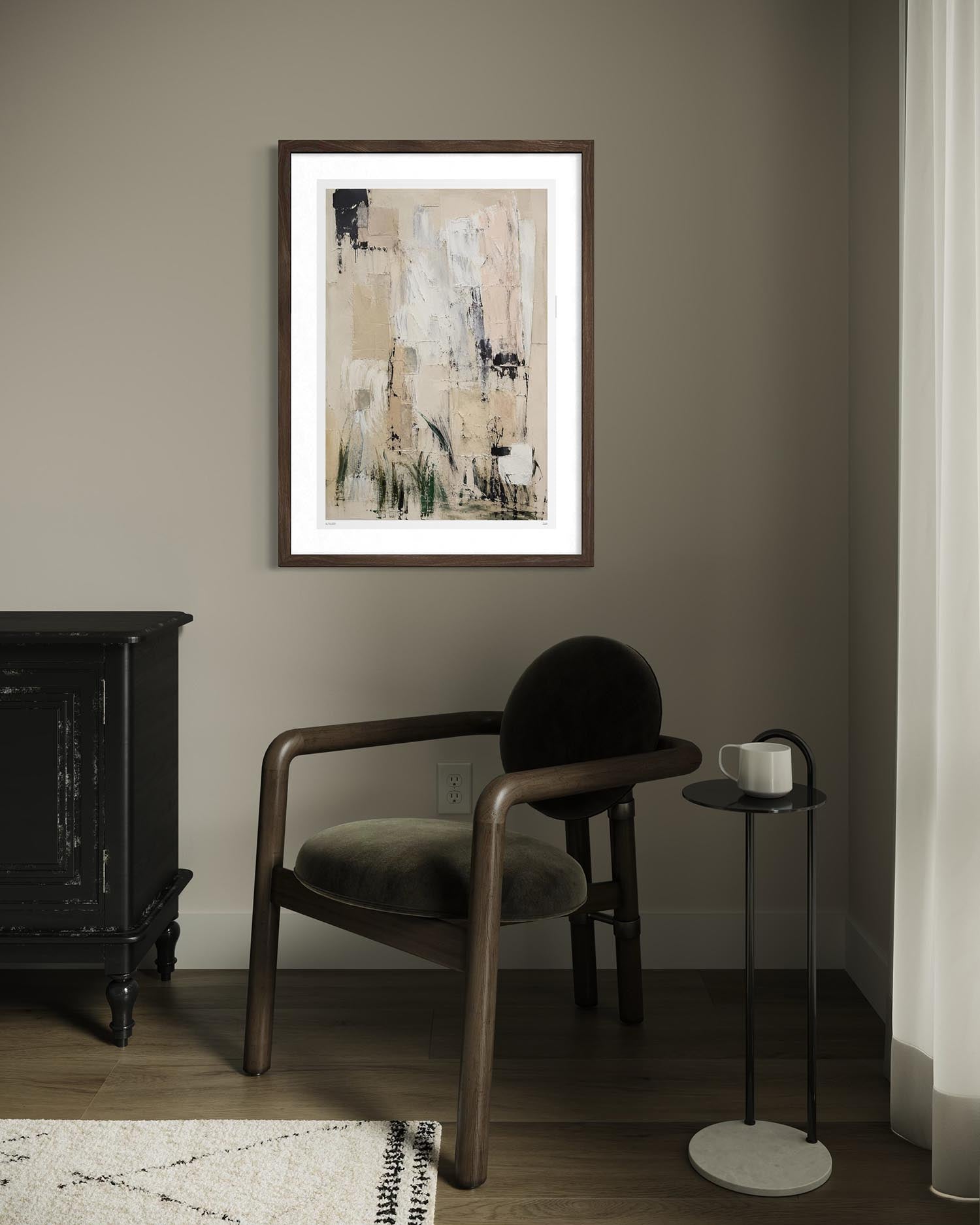 A framed abstract art print with beige, black, and white brushstrokes hangs on a taupe-gray wall in a styled room setting with a wooden armchair, a side table with a white mug, and a dark cabinet.