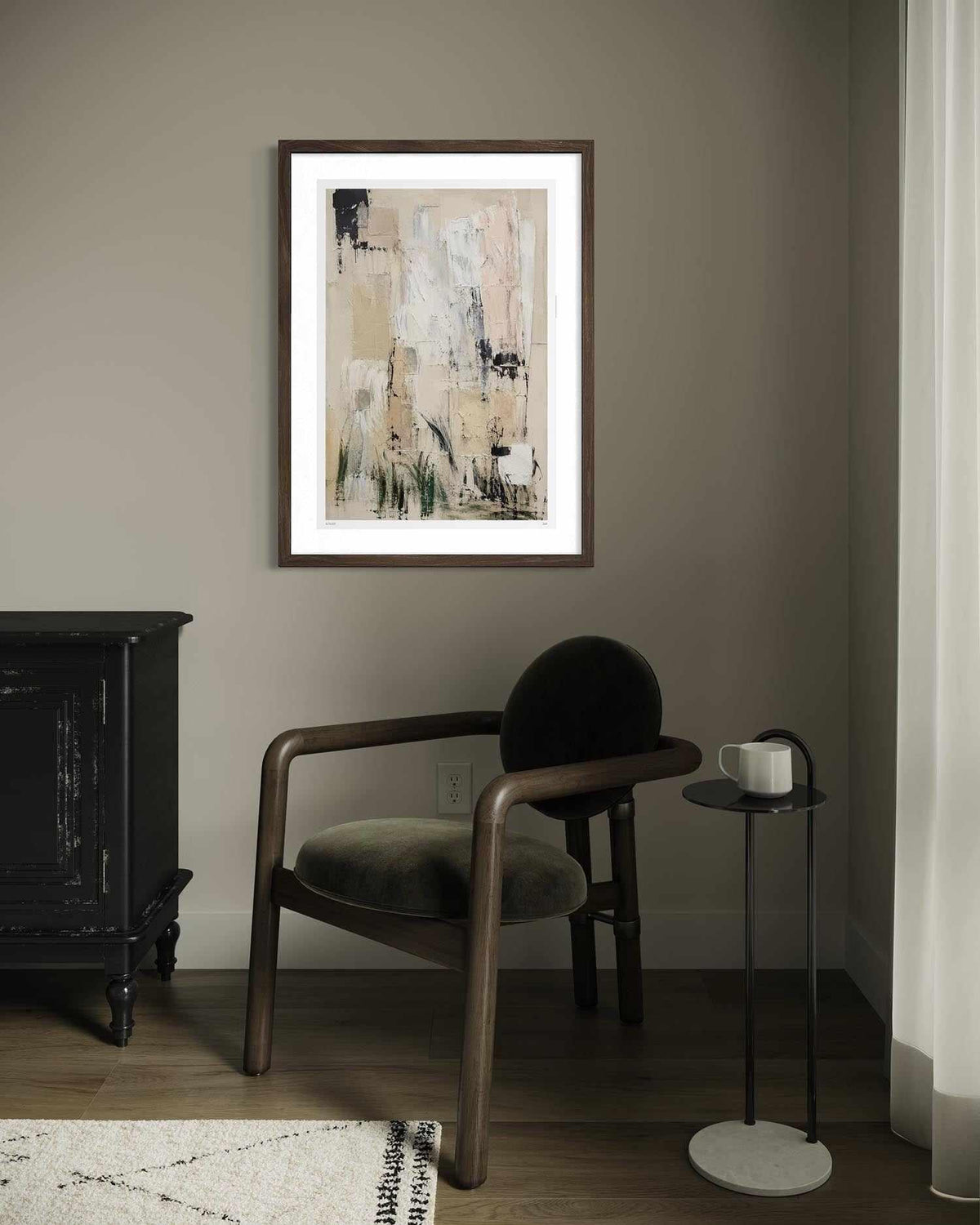 A framed abstract art print with beige, black, and white brushstrokes hangs on a taupe-gray wall in a styled room setting with a wooden armchair, a side table with a white mug, and a dark cabinet.