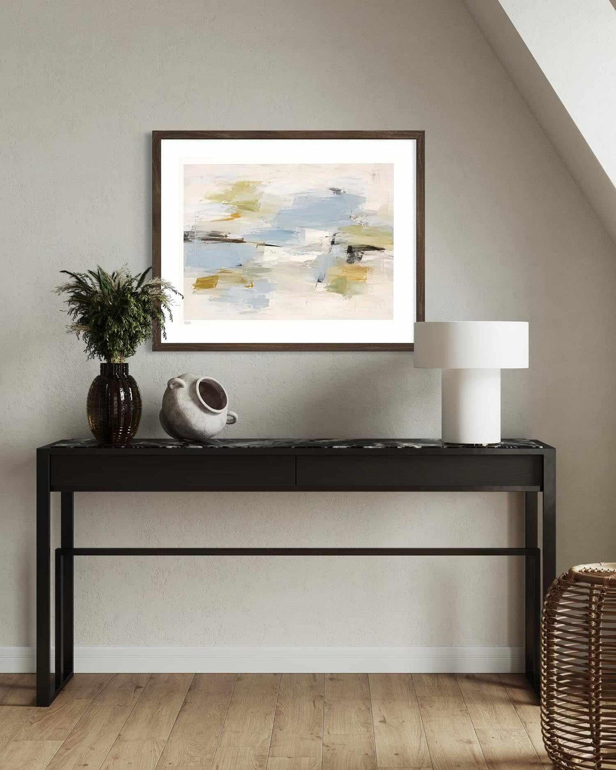 An abstract framed art print with a white mat and dark brown frame, displayed on a black console table in a modern room setting with a vase, ceramic pot, and lamp.