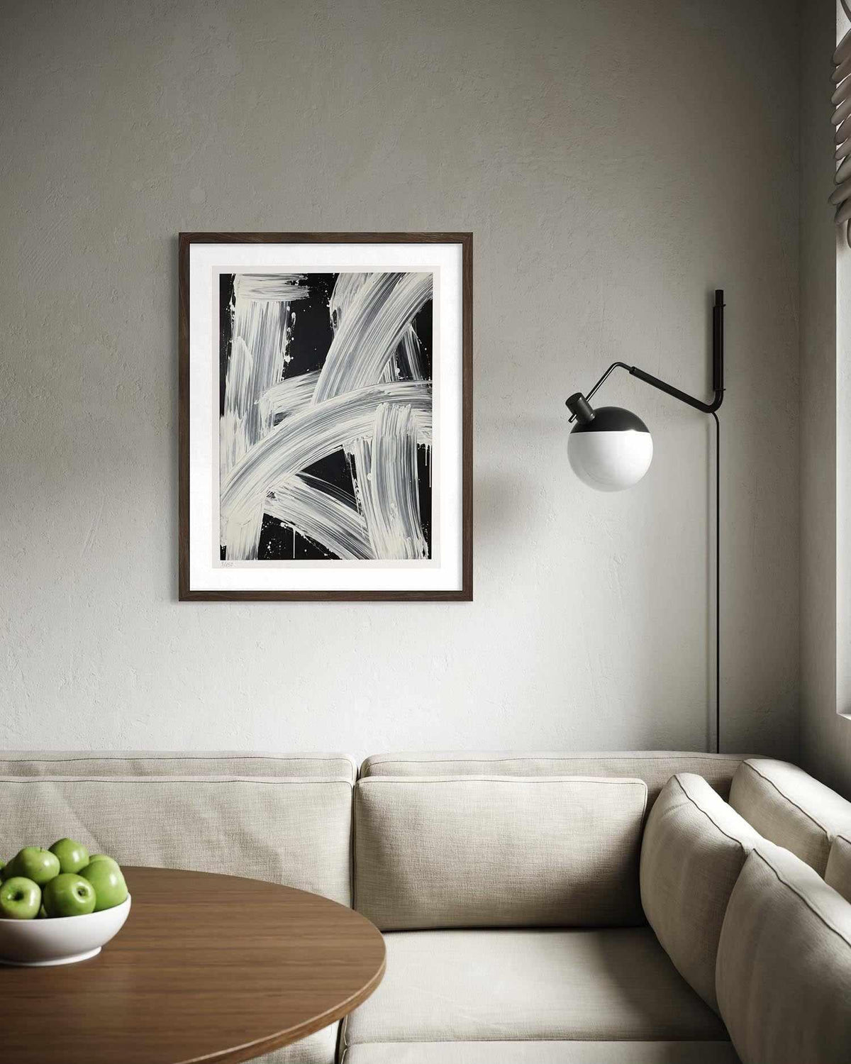 A framed abstract black and white art print hangs on a textured gray wall above a beige sectional sofa, with a wooden side table holding green apples in the foreground and a black floor lamp to the right.