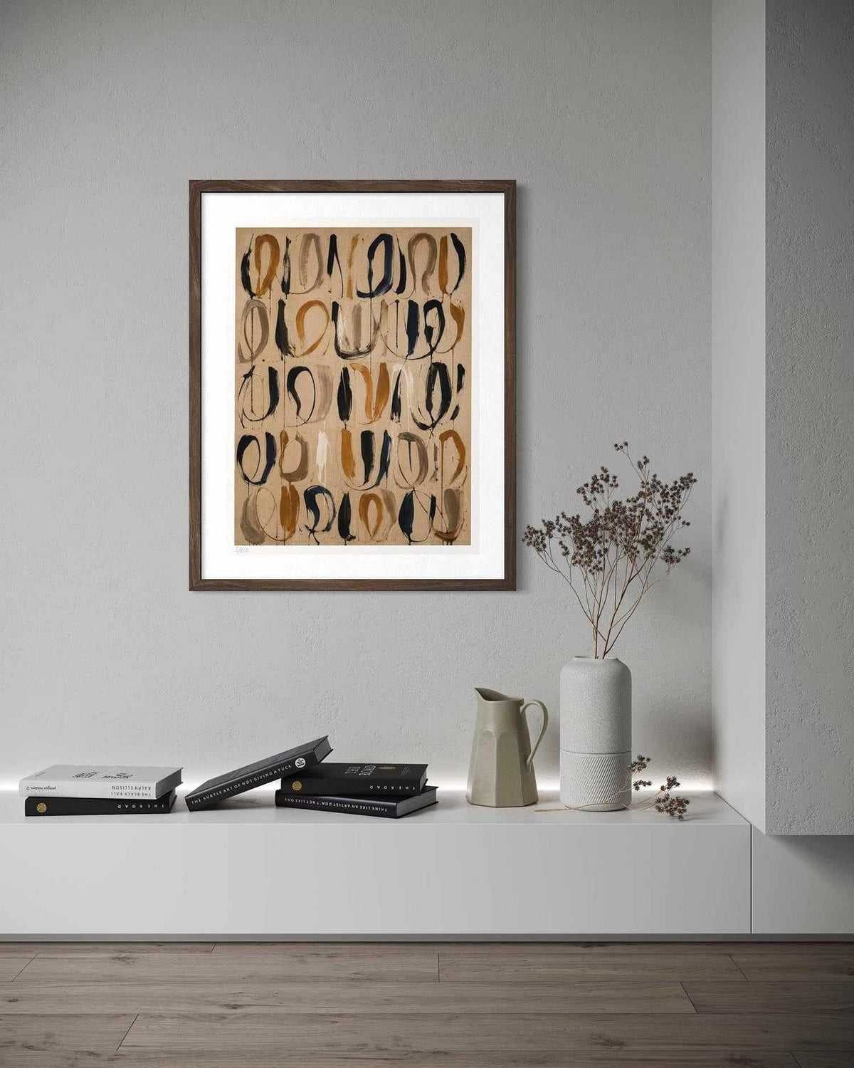 A framed abstract art print with a white mat and dark brown frame, hanging on a light gray wall above a white console table styled with books, a ceramic pitcher, and a vase with dried branches.
