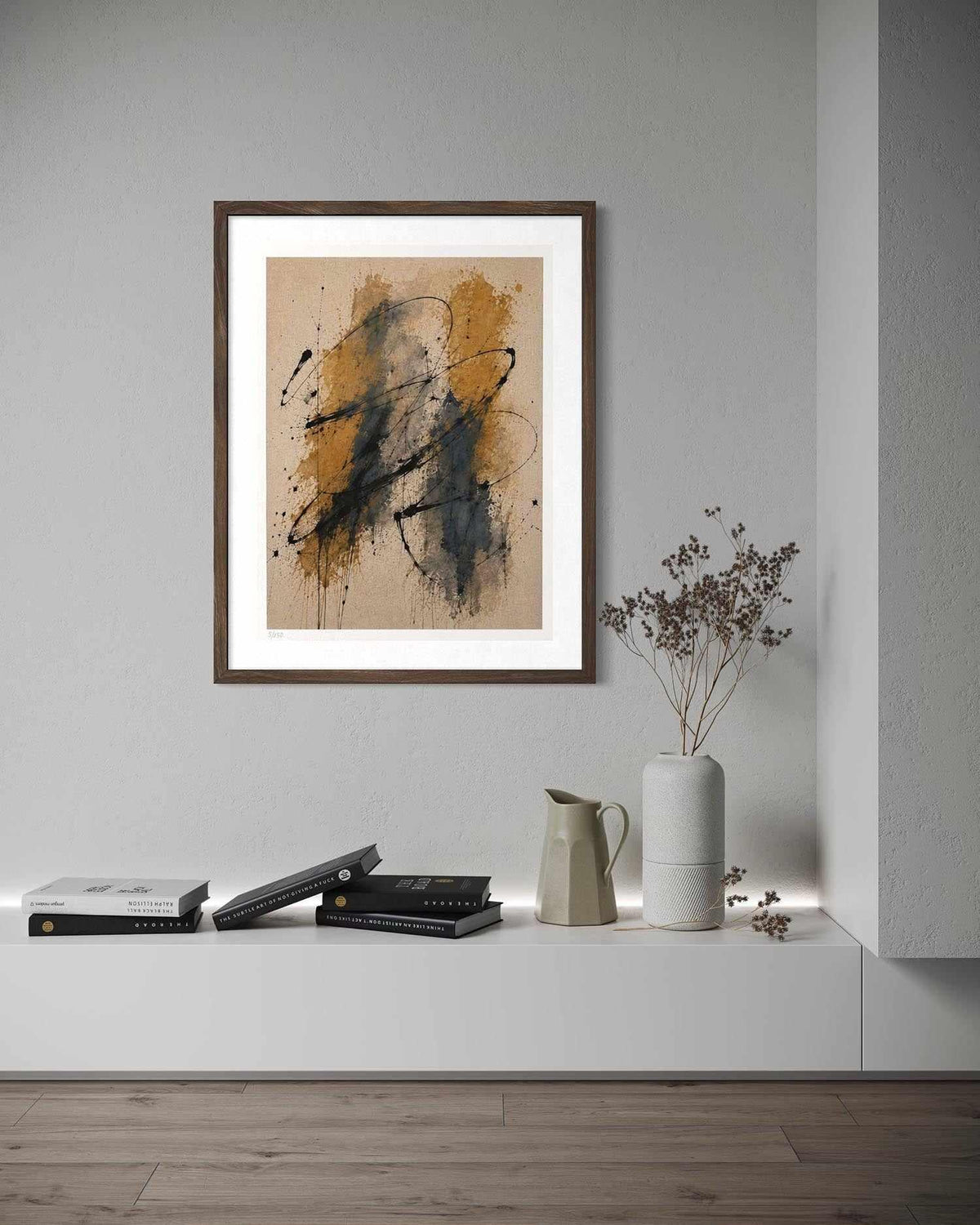 A framed abstract art print with black and ochre brushstrokes on a beige background, displayed on a light gray wall above a white console table with books, a ceramic pitcher, and a vase with dried branches.