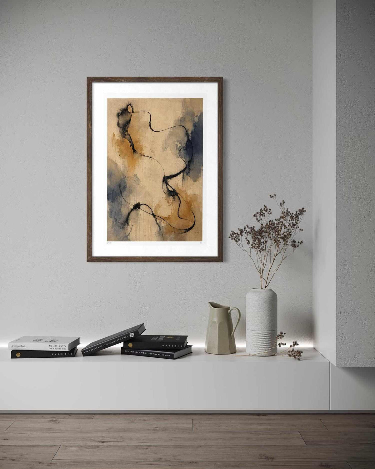 A framed abstract art print with a white mat and dark brown frame, hanging on a light gray wall above a white shelf with books, a ceramic pitcher, and dried flowers.