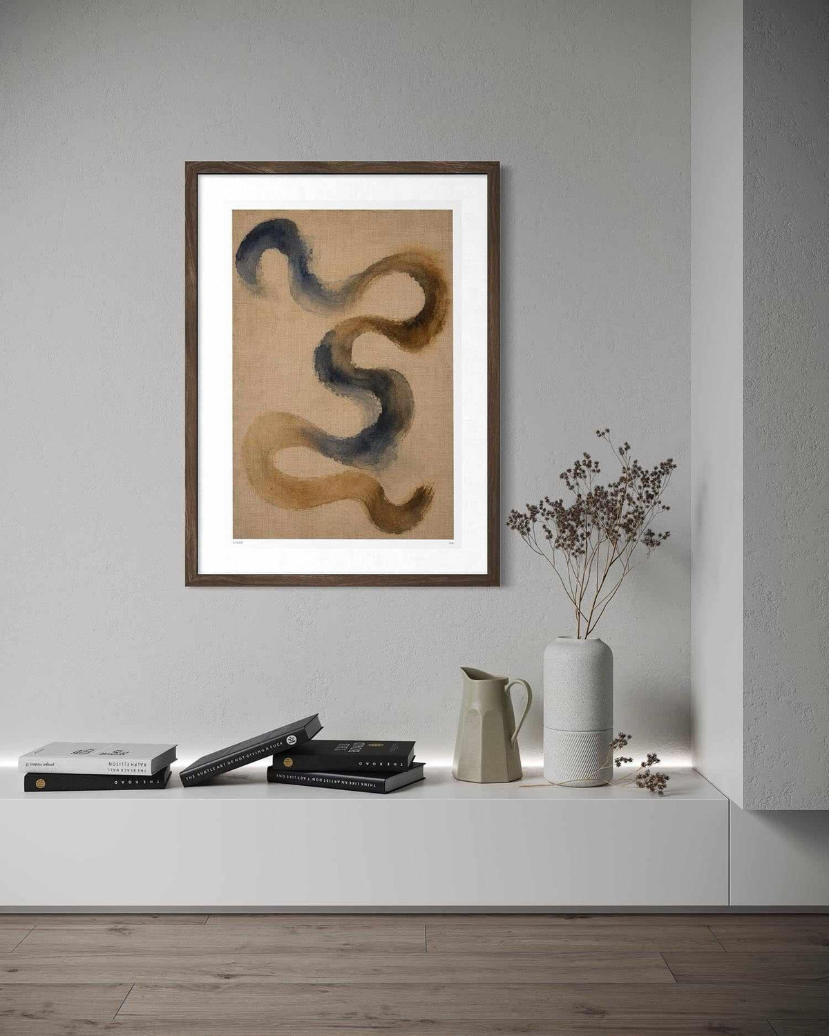 Framed abstract wall art hanging on a light gray wall above a white shelf with books, a ceramic pitcher, and a vase with dried branches. The artwork shows flowing curved brushstrokes in tan, brown, and dark blue-gray on a beige background.