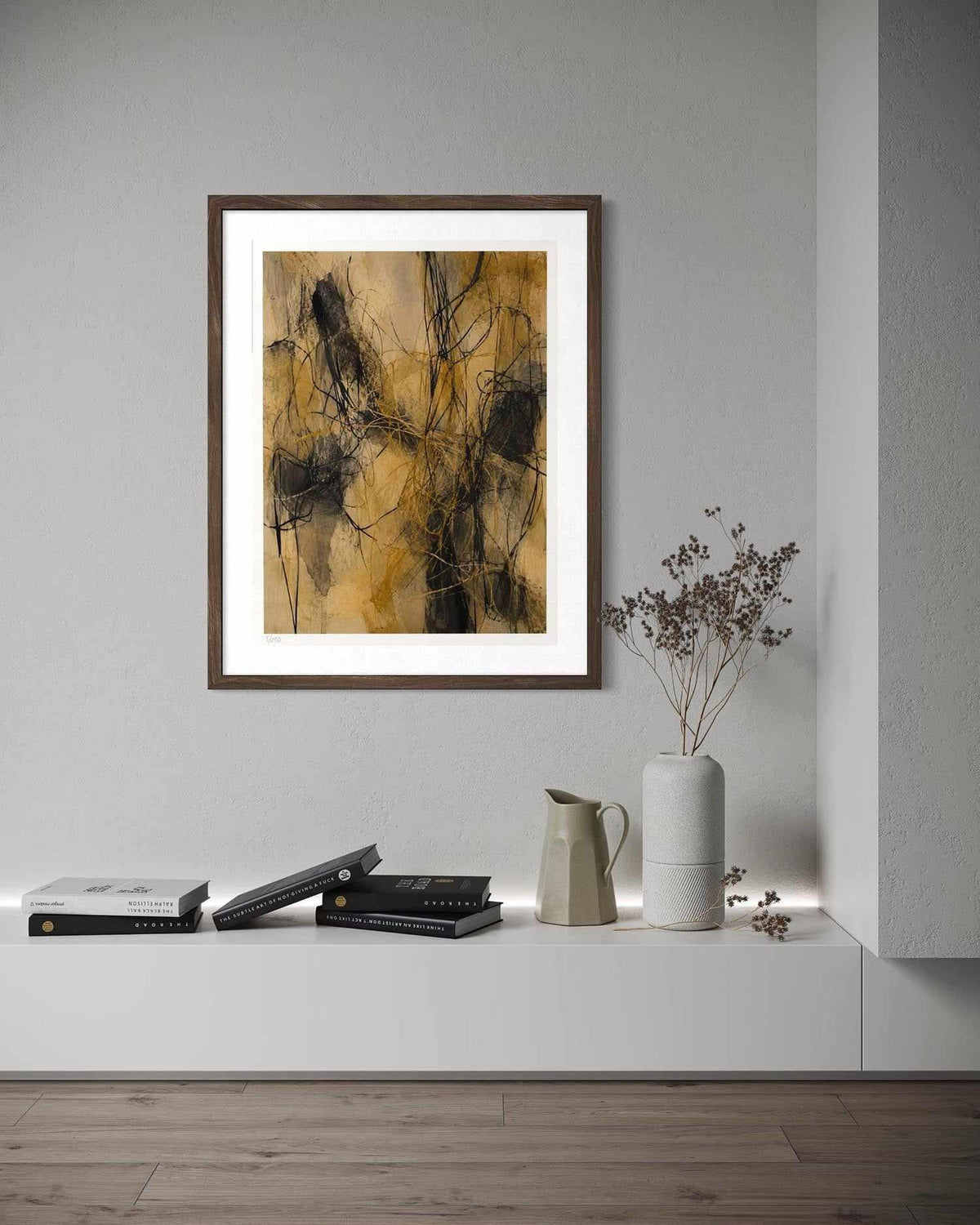 Framed abstract wall art hanging on a light gray wall above a white console shelf with books, a ceramic pitcher, and a vase with dried stems.