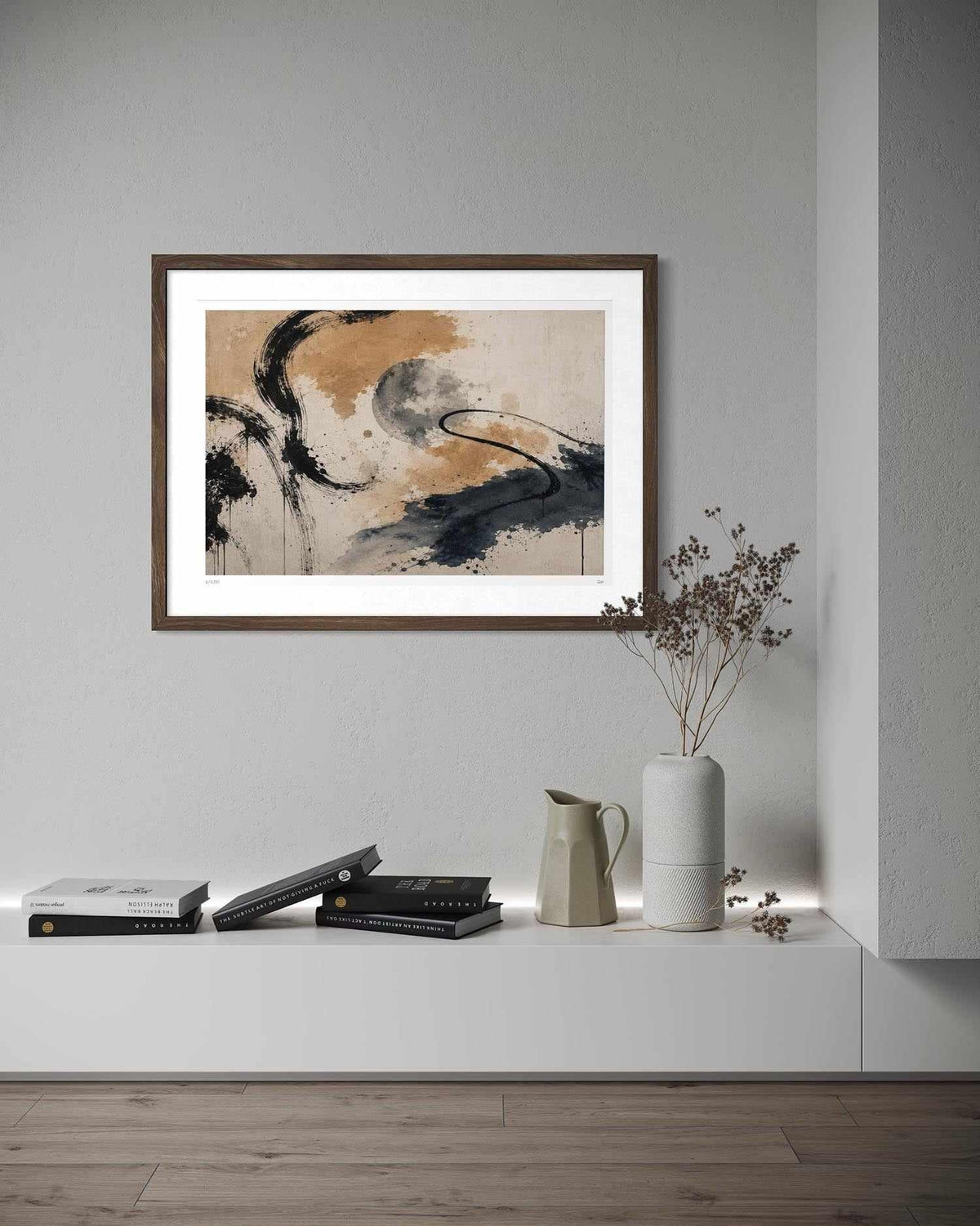 A framed abstract art print with black, gray, and beige brushstrokes on a white mat, hanging on a light gray wall above a white shelf with books, a ceramic pitcher, and a vase with dried branches.
