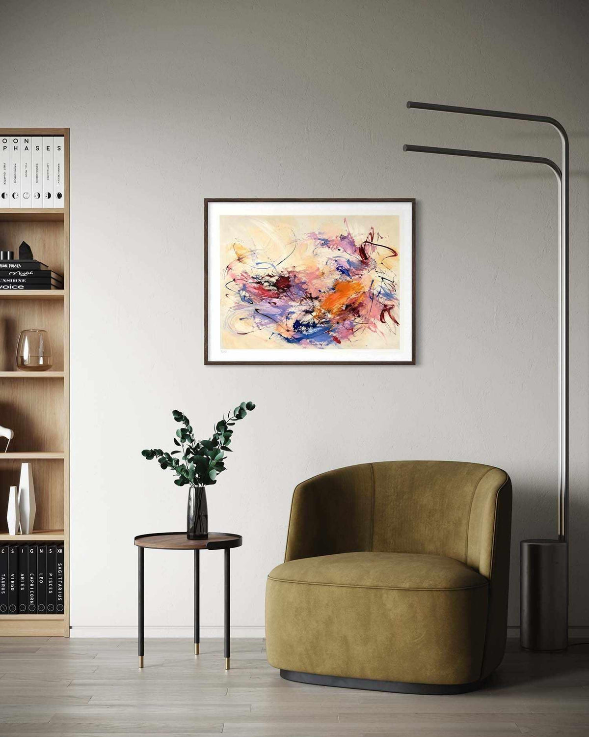 A framed abstract art print with vibrant splashes of orange, purple, pink, and blue on a cream background, displayed on a light gray wall in a modern living room setting with a green armchair and a bookshelf.