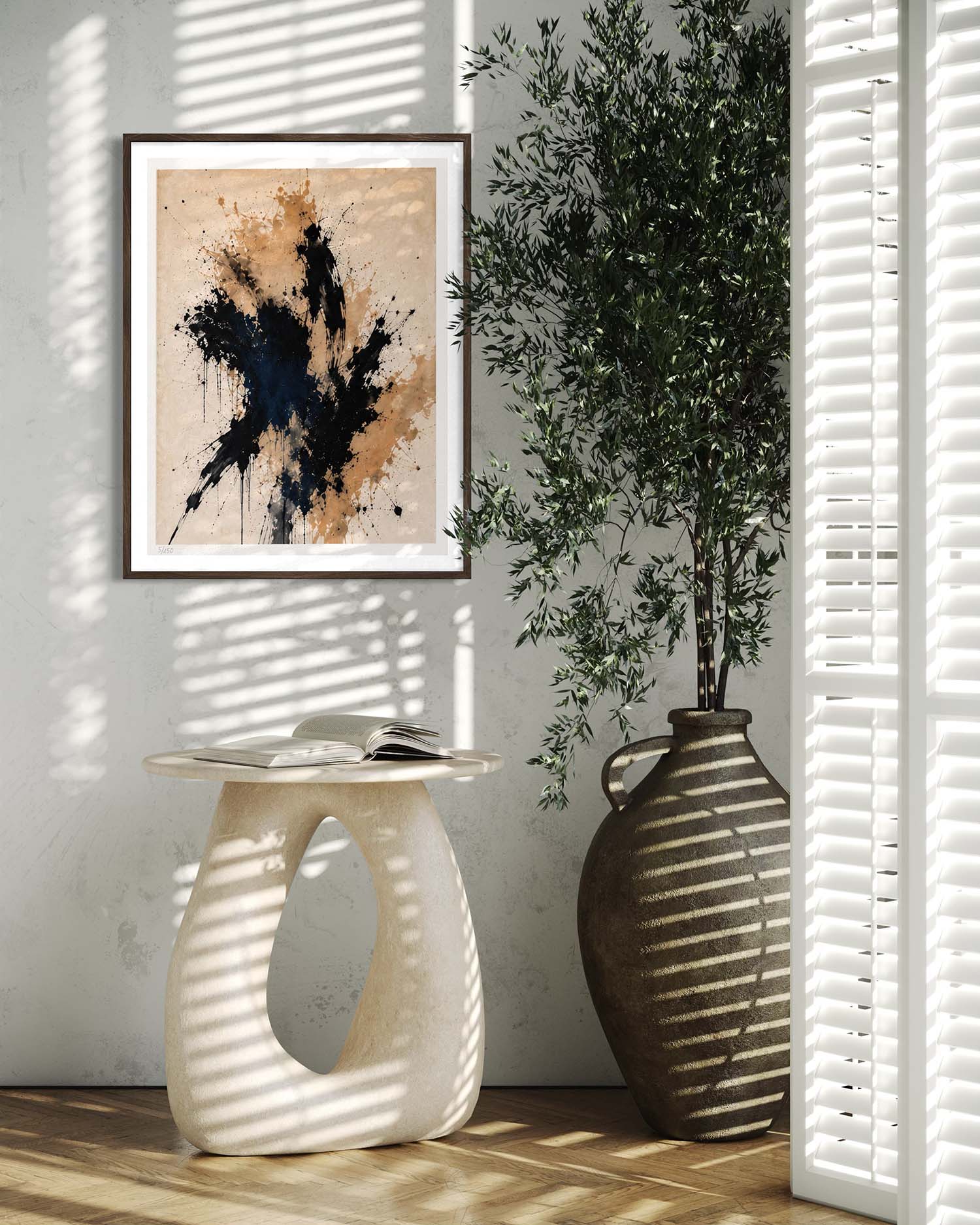 Framed abstract wall art hanging on a light wall in a styled interior, with a sculptural side table, open book, large vase, and potted plant in soft window-shadow lighting.