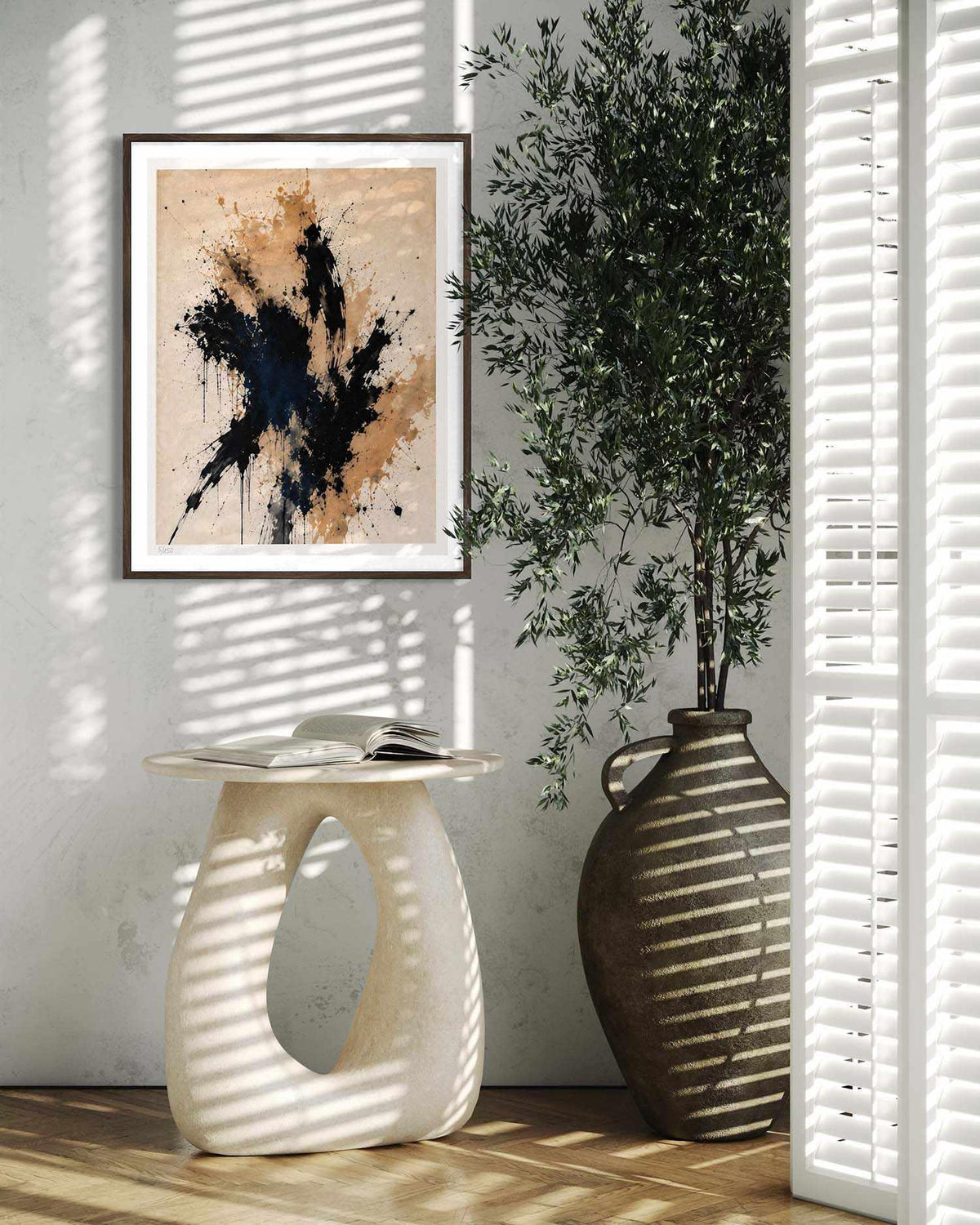 Framed abstract wall art hanging on a light wall in a styled interior, with a sculptural side table, open book, large vase, and potted plant in soft window-shadow lighting.