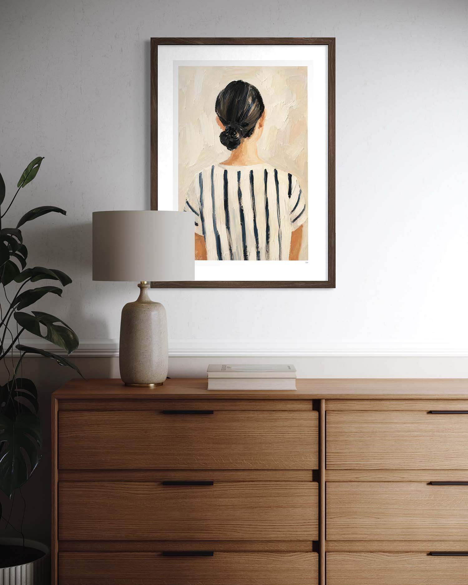 A framed art print depicting a woman from behind wearing a black and white striped top, styled on a white wall above a wooden dresser with a lamp and plant.
