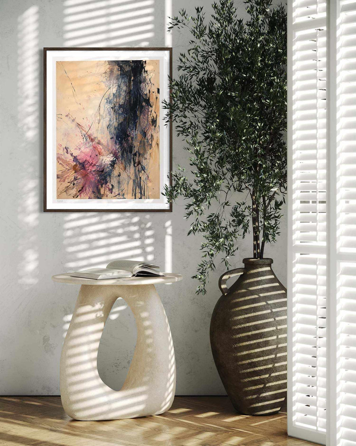 A framed abstract art print hangs on a light gray wall in a styled interior scene with a sculptural side table, a large vase with a tree, and window blinds casting striped shadows.
