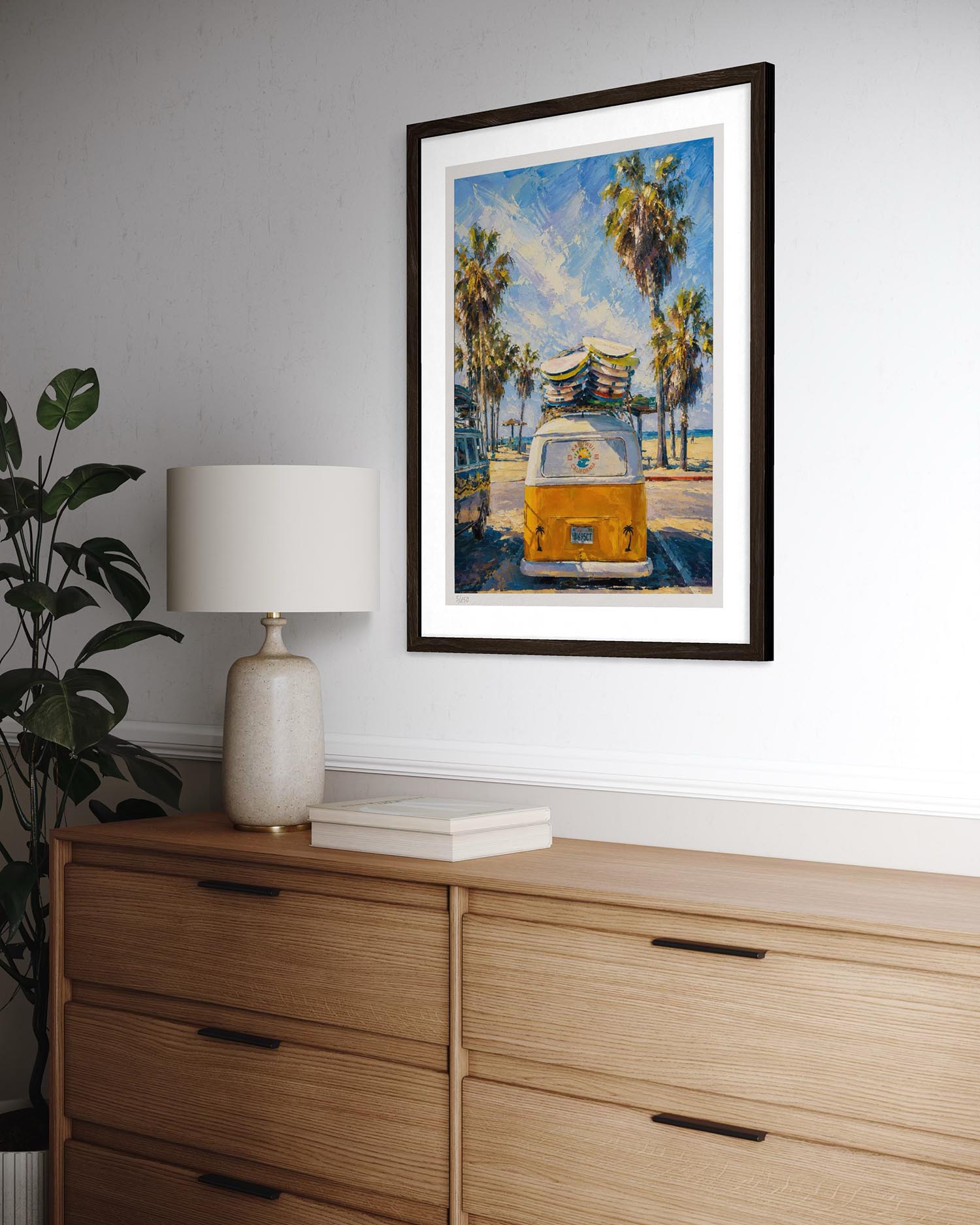 A framed art print depicting a yellow VW bus on a beach with palm trees, styled on a white wall above a wooden dresser with a lamp and plant.