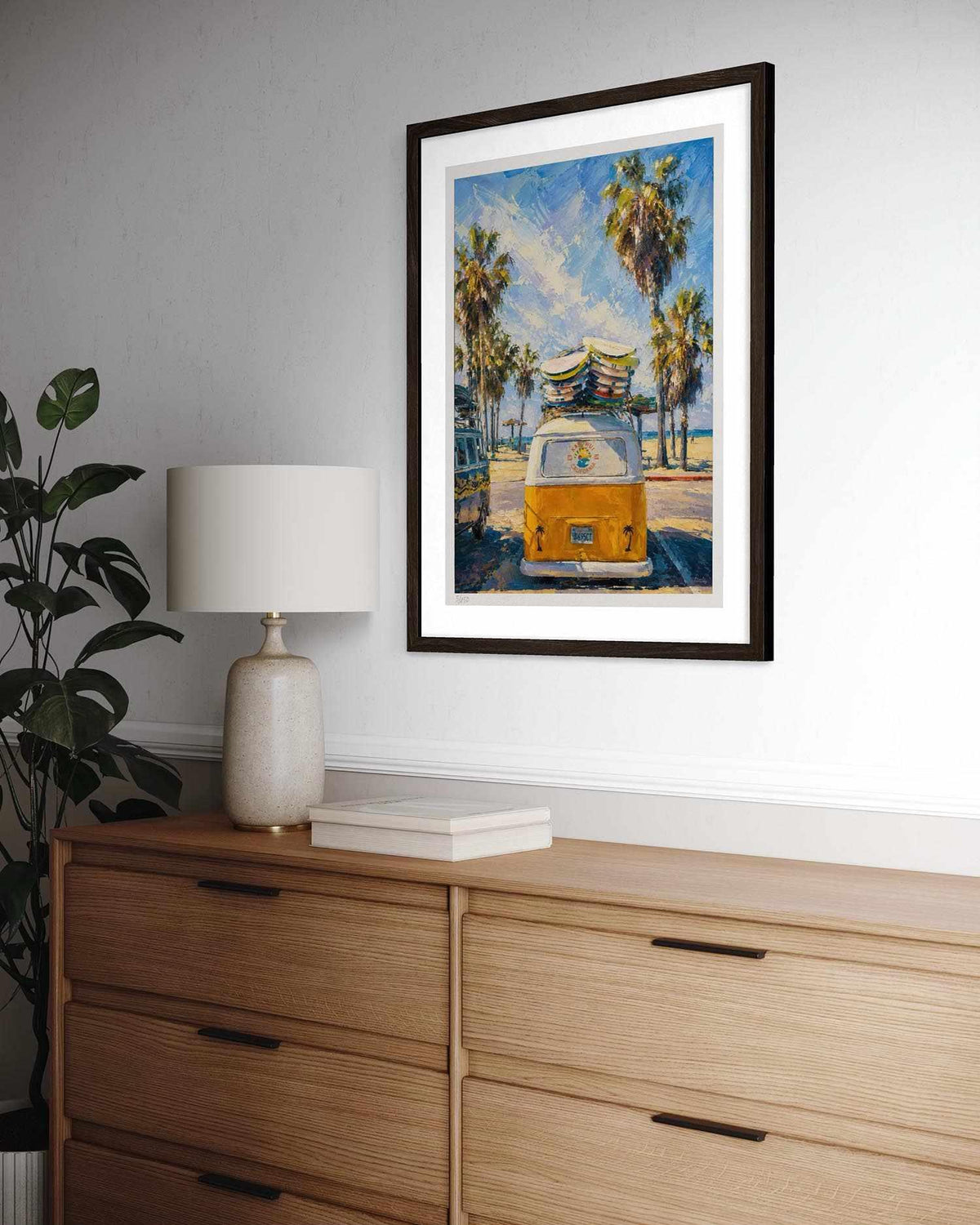 A framed art print depicting a yellow VW bus on a beach with palm trees, styled on a white wall above a wooden dresser with a lamp and plant.