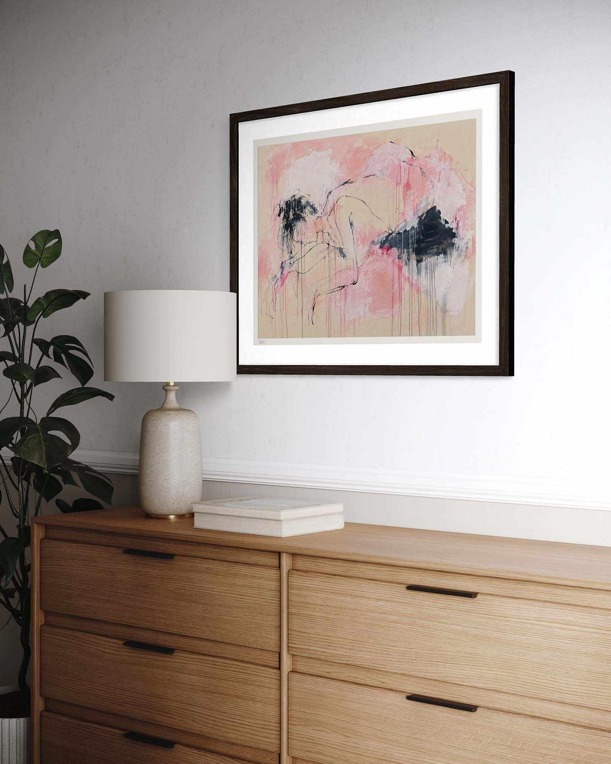 A framed abstract art print featuring pink, beige, and black brushstrokes, displayed on a white wall above a light wood dresser with a lamp and plant.