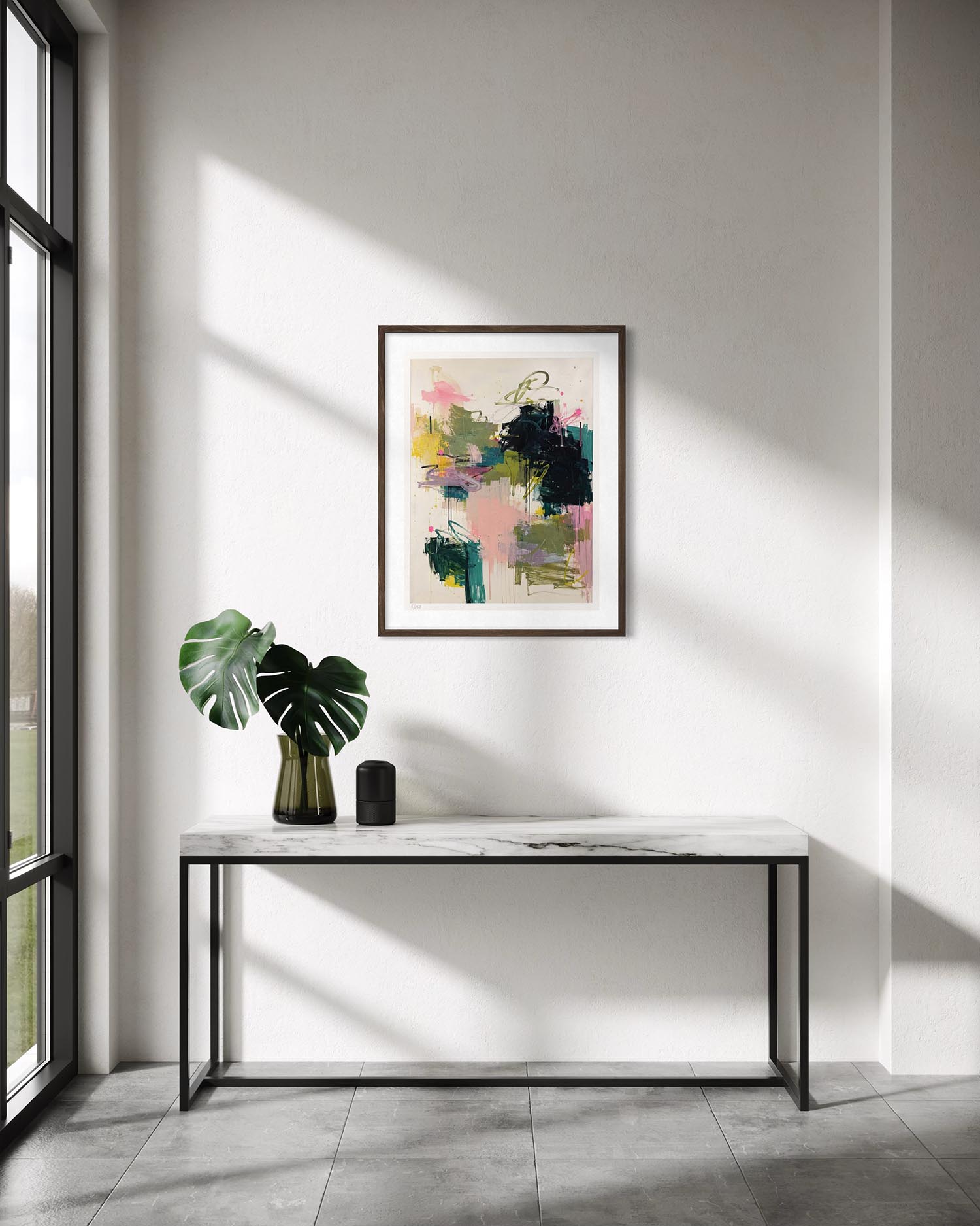 A framed abstract art print with pink, green, yellow, and black brushstrokes hangs on a white wall above a marble-top console table with a green plant and a small black speaker in a sunlit room.