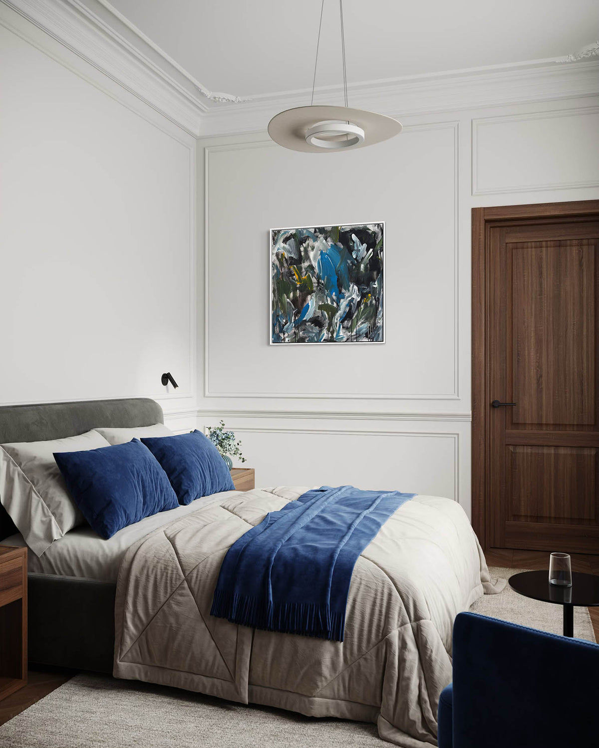 A styled bedroom interior featuring a bed dressed in beige bedding with two dark blue pillows and a blue throw blanket, set against white paneled walls with a framed abstract painting and a dark wood door.