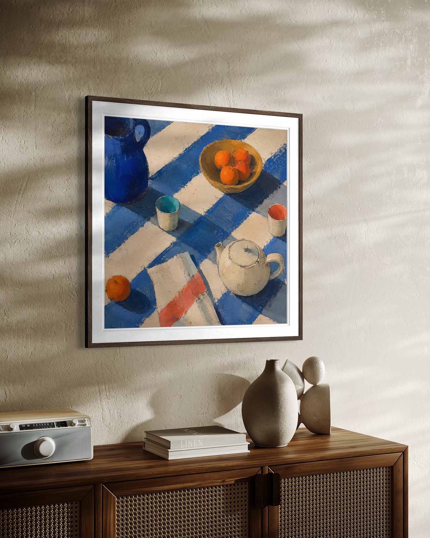 A framed still life painting hanging on a textured beige wall above a wooden sideboard. The artwork depicts a blue jug, a bowl of oranges, a teapot, and other table objects on a blue-and-white checkered cloth.
